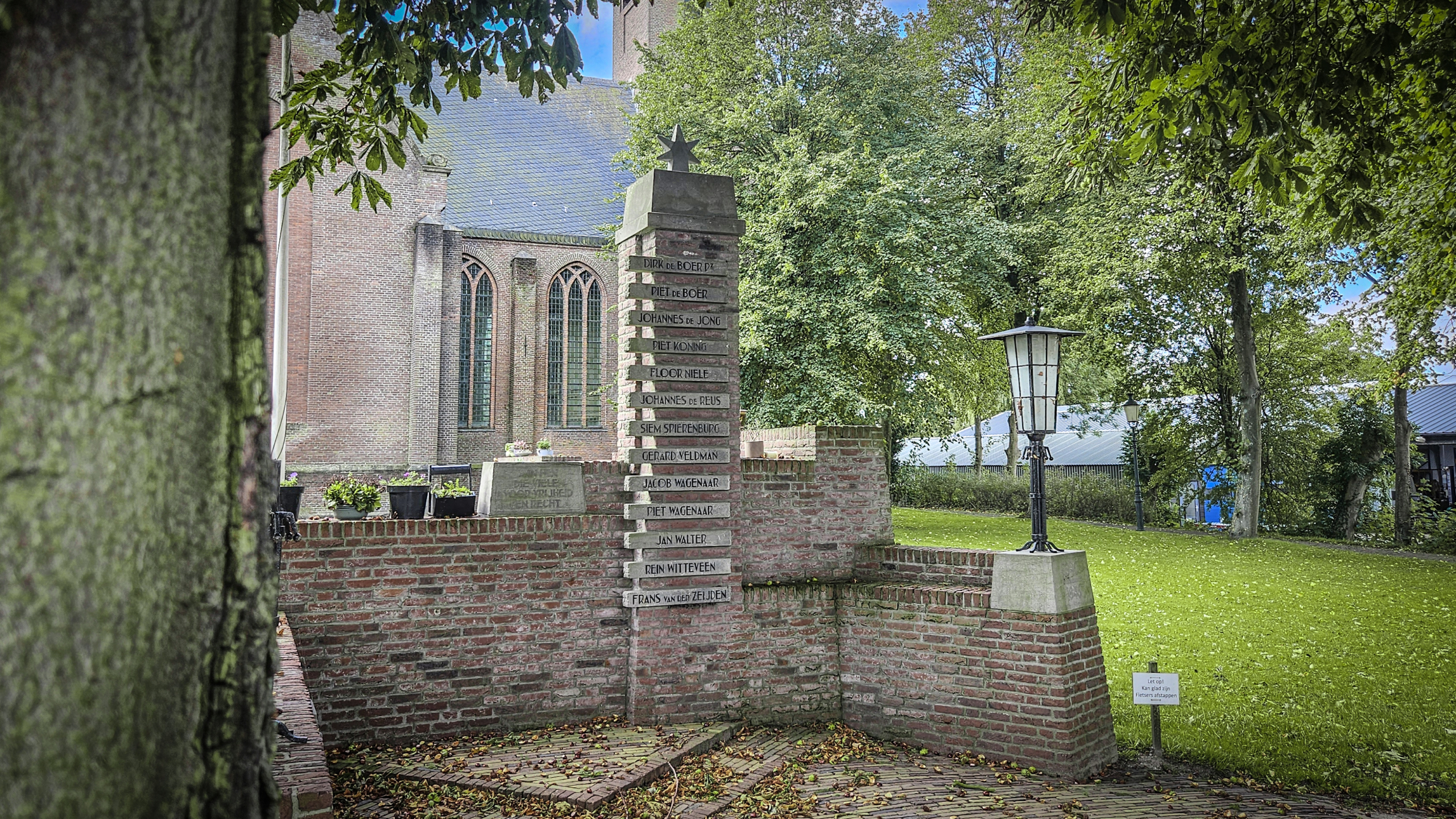 Een monument met namen van slachtoffers staat naast een historische kerk, omringd door groen en een lantaarnpaal in een rustige, landelijke omgeving.