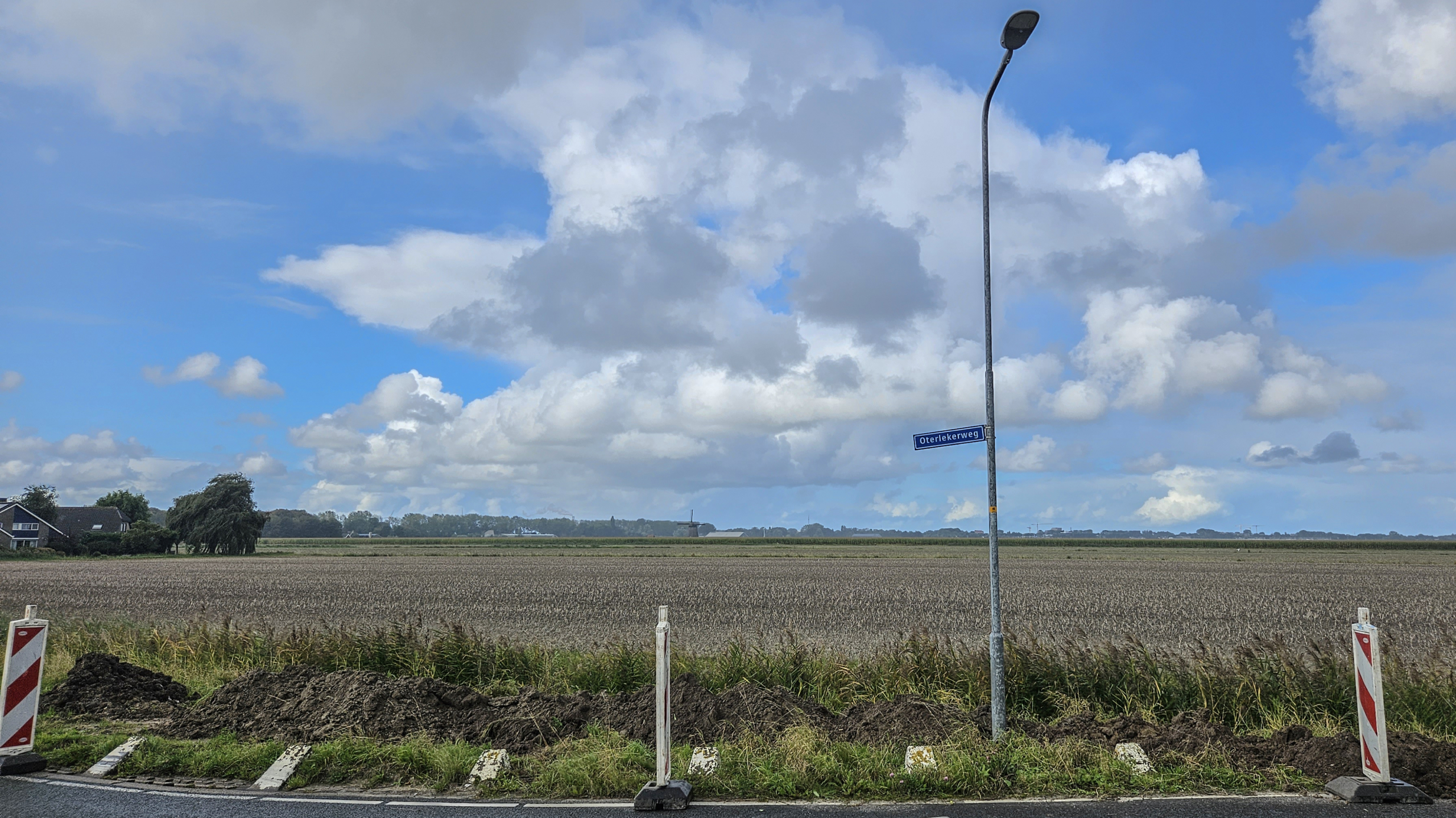 Een landelijk landschap met een uitgestrekte akker onder een blauwe hemel met wolken, naast een straatlantaarn met het bordje 'Oterlekerweg' en wegwerkzaamheden aangegeven door borden.