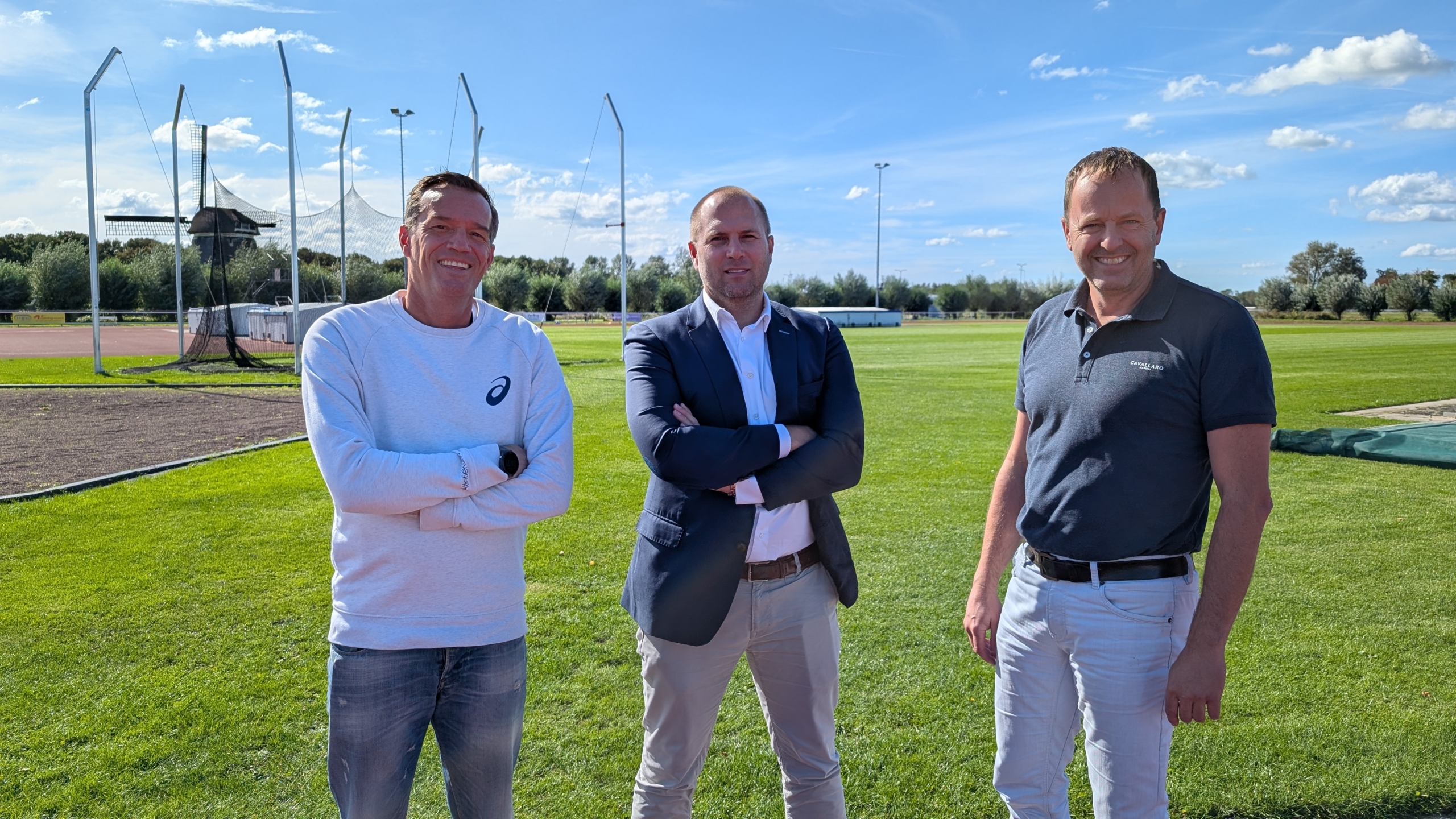 Drie mannen staan op een groene sportveld met een windmolen op de achtergrond, onder een blauwe lucht met wolken.