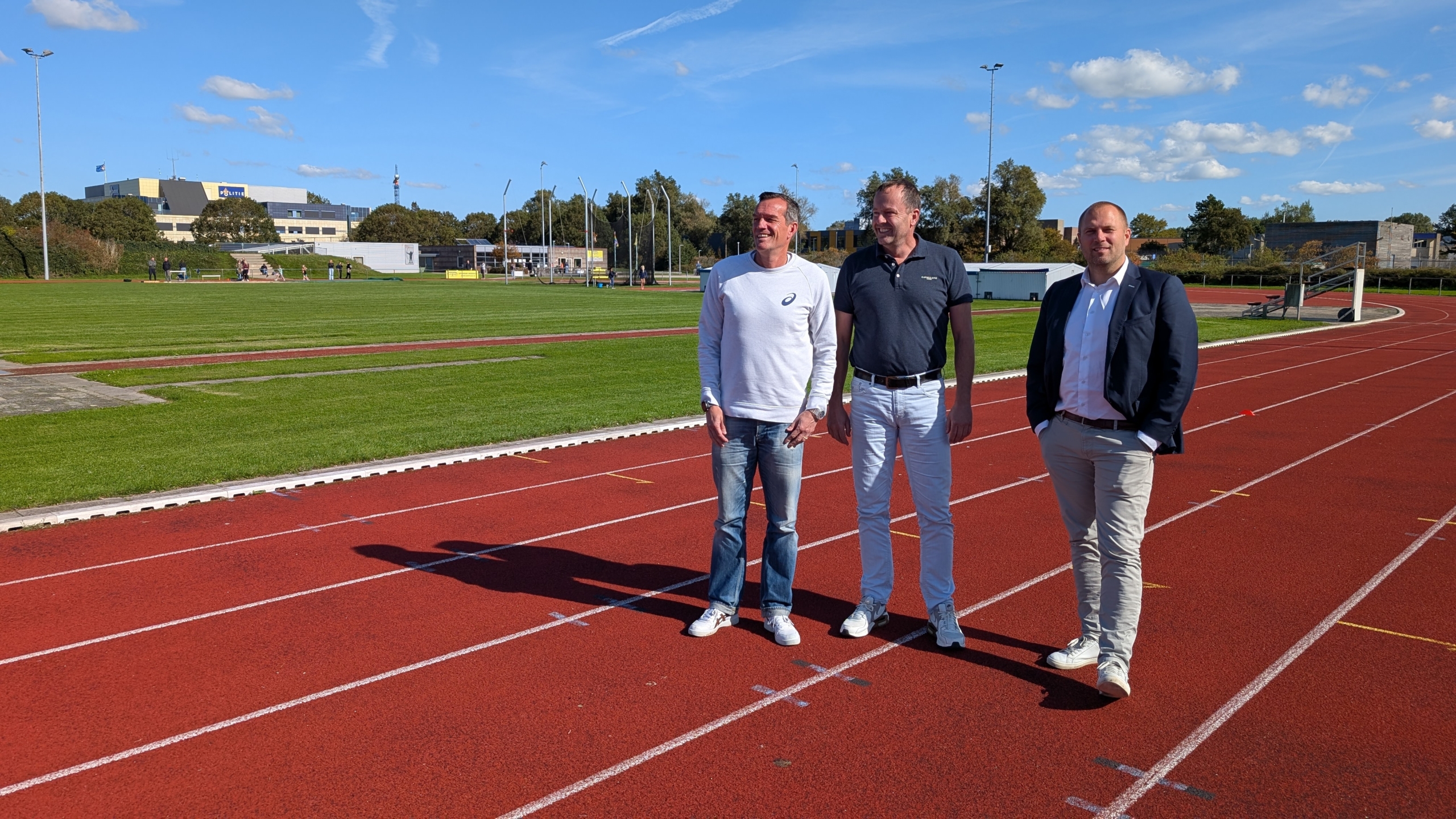 Drie mannen staan op een atletiekbaan met een groene grasmat en een gebouw in de achtergrond op een zonnige dag.