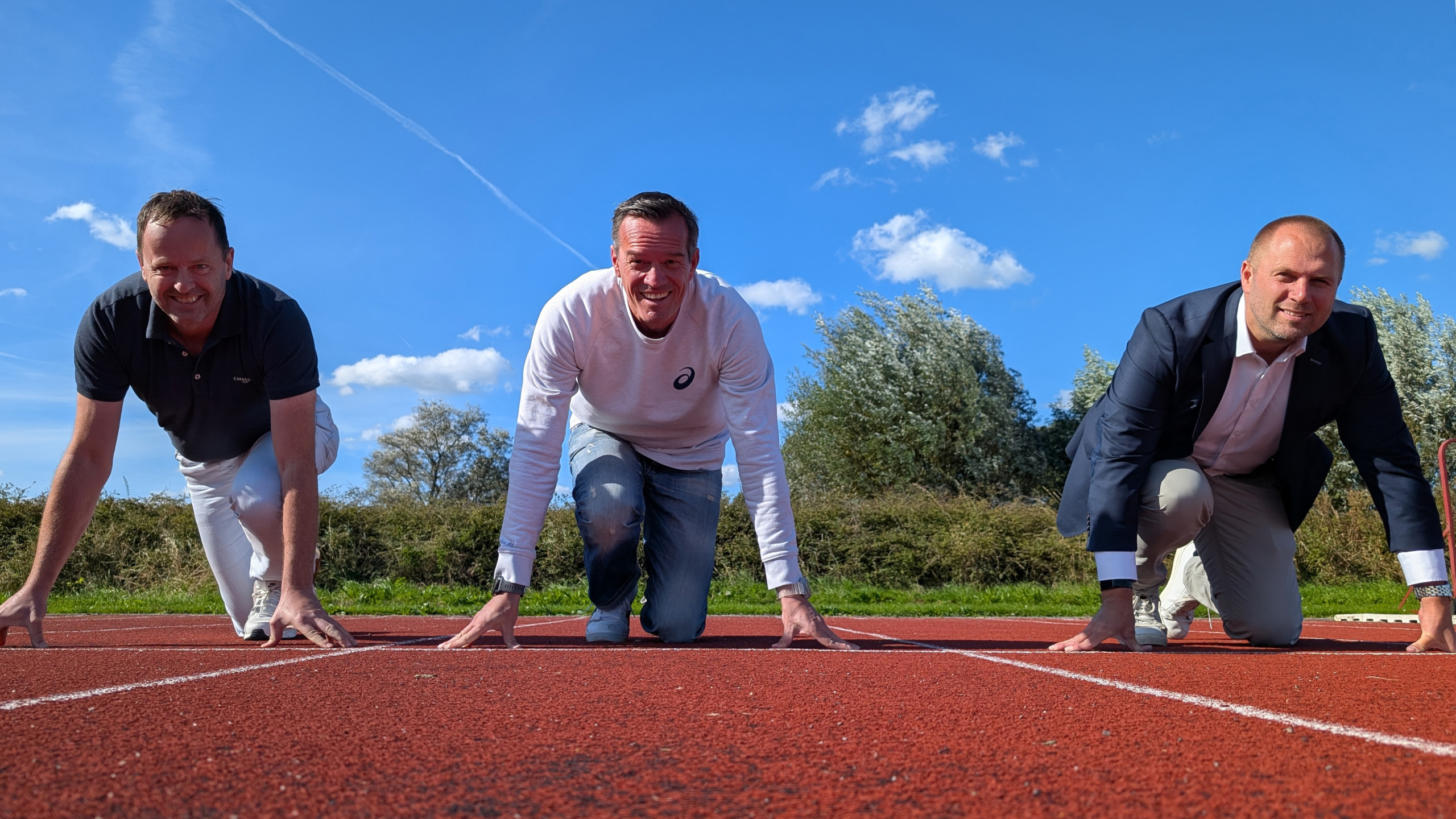 Drie mannen staan in startpositie op een atletiekbaan, met een heldere blauwe lucht op de achtergrond.