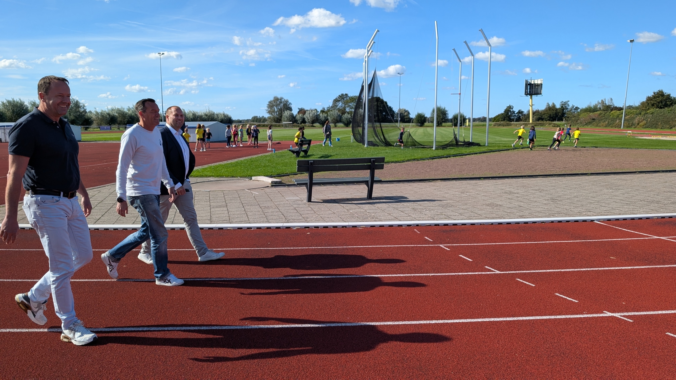 Drie mensen lopen op een atletiekbaan naast een grasveld, waar kinderen aan het sporten zijn, op een zonnige dag met een blauwe lucht vol wolken.