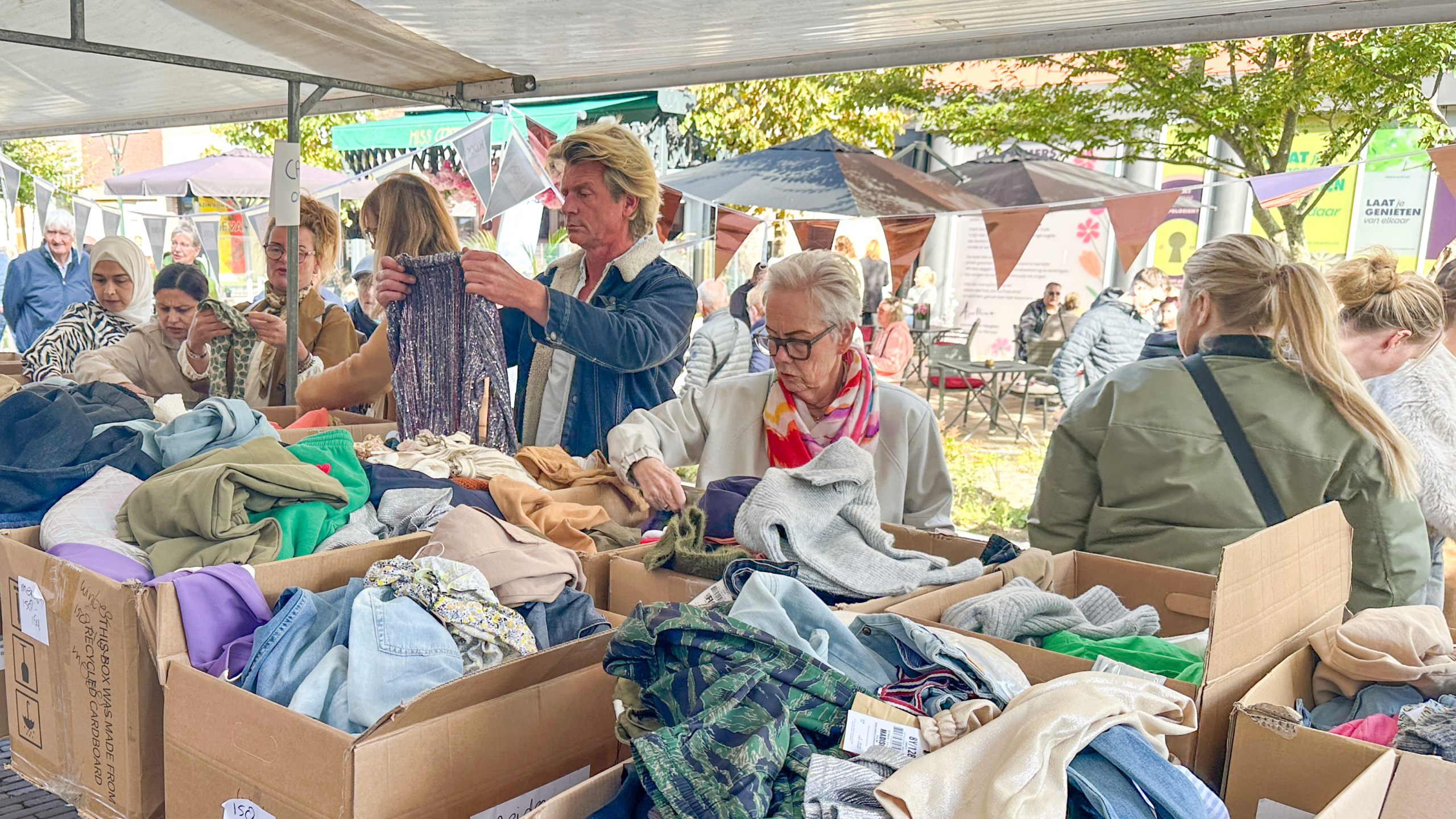 Mensen bladeren door kleding in kartonnen dozen op een openluchtmarkt, omringd door andere bezoekers en tafels met parasols in de achtergrond.