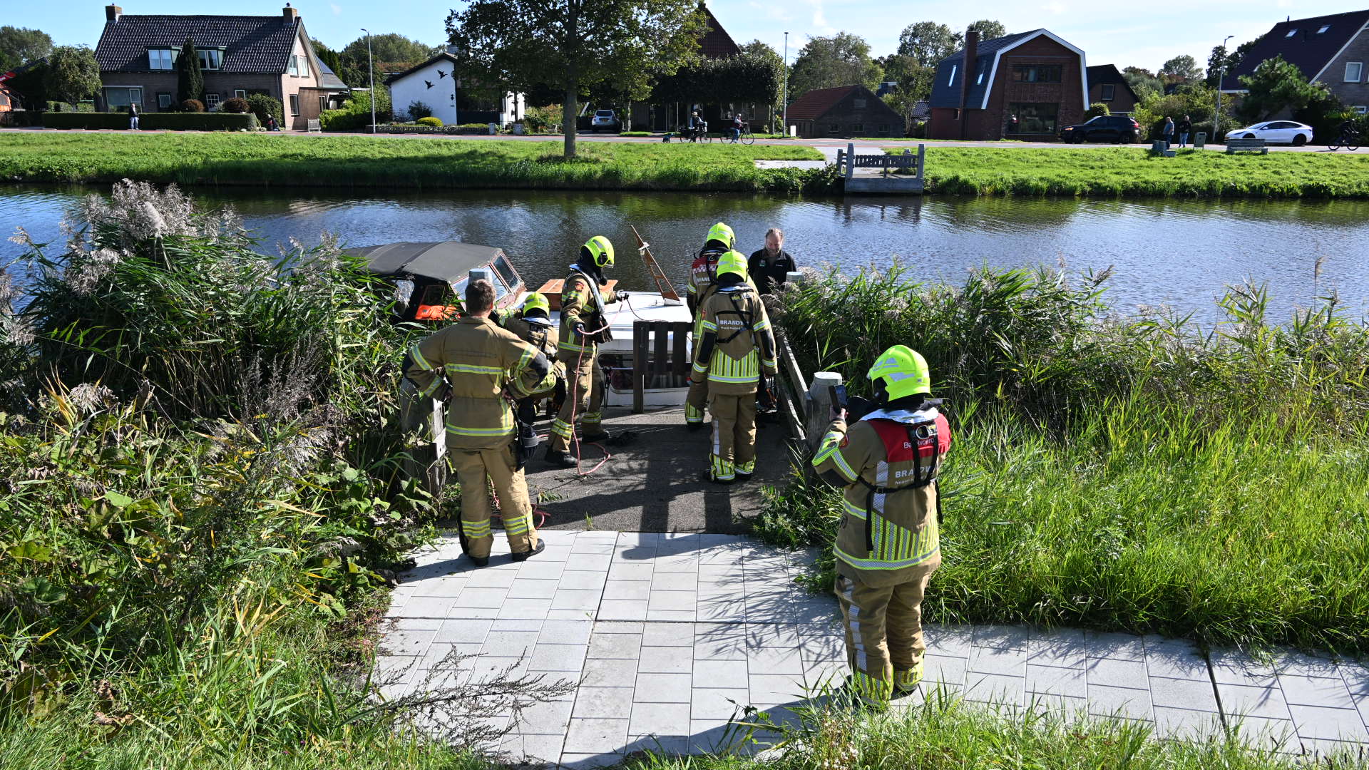 Brandweerlieden in gele veiligheidskleding staan bij een kleine boot aan de oever van een rivier, omgeven door hoge grassen, met huizen en een park op de achtergrond.