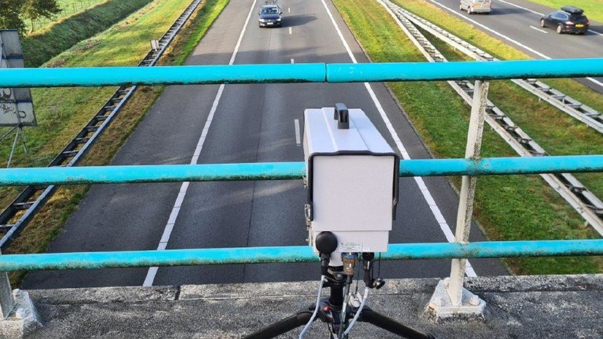 Snelheidscamera op statief boven een viaduct dat uitkijkt op een snelweg met passerende auto's.