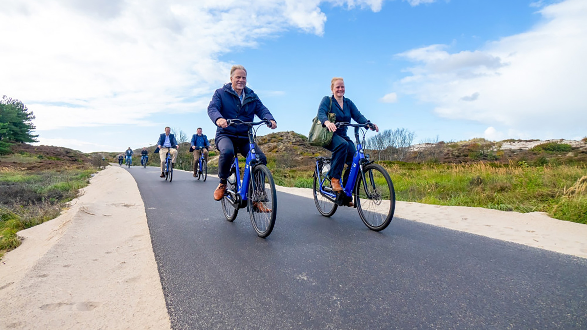 Mensen fietsen op een zonnige dag over een geasfalteerd pad in een natuurlijke omgeving met gras en zandduinen.