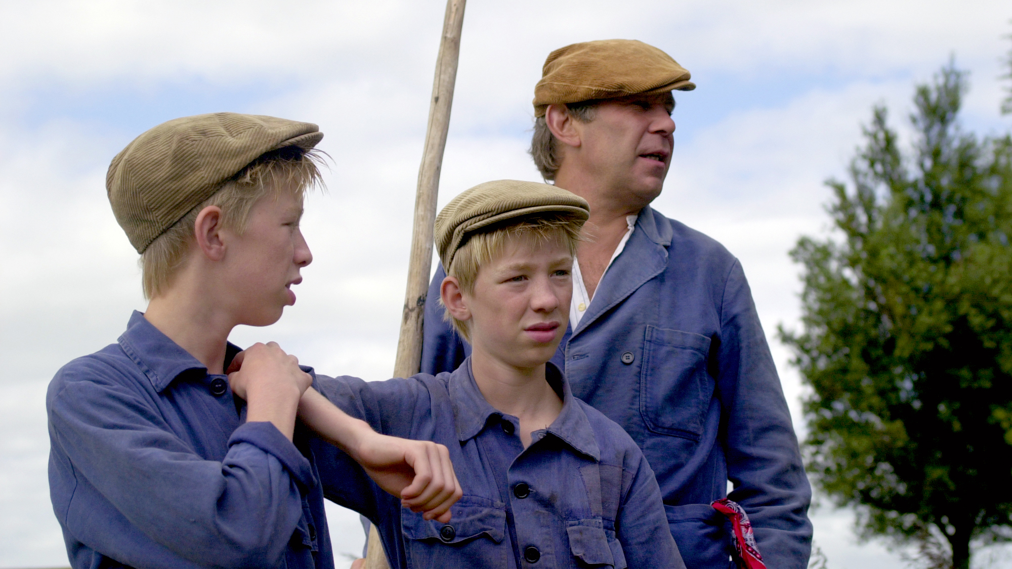 Twee jongens en een man dragen blauwe overalls en bruine petten, terwijl ze buiten staan met een stok bij bewolkte hemel.