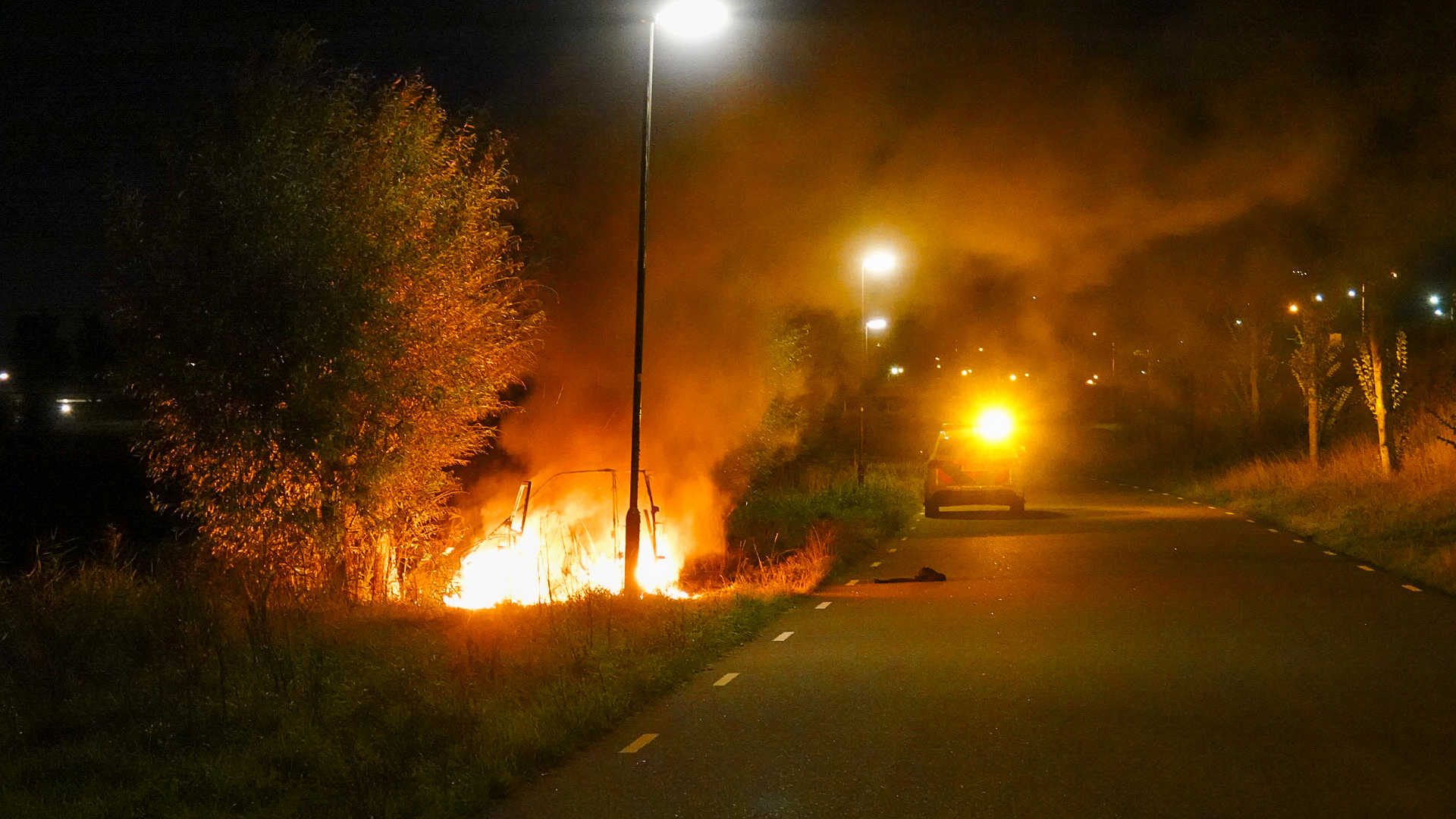 Brandende auto langs een verlaten weg 's nachts met rook en een gele waarschuwingslamp in de verte.