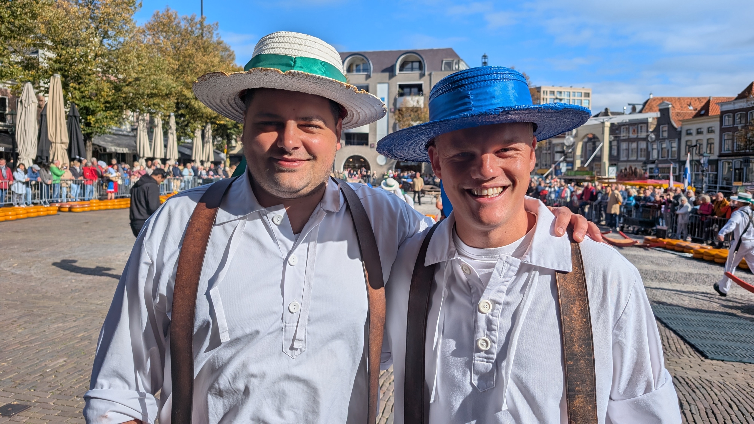 Twee mannen in traditionele witte kleding en strohoeden, één met een groene band en de ander met een blauwe band, poseren op een drukke markt met toeschouwers en historische gebouwen op de achtergrond.