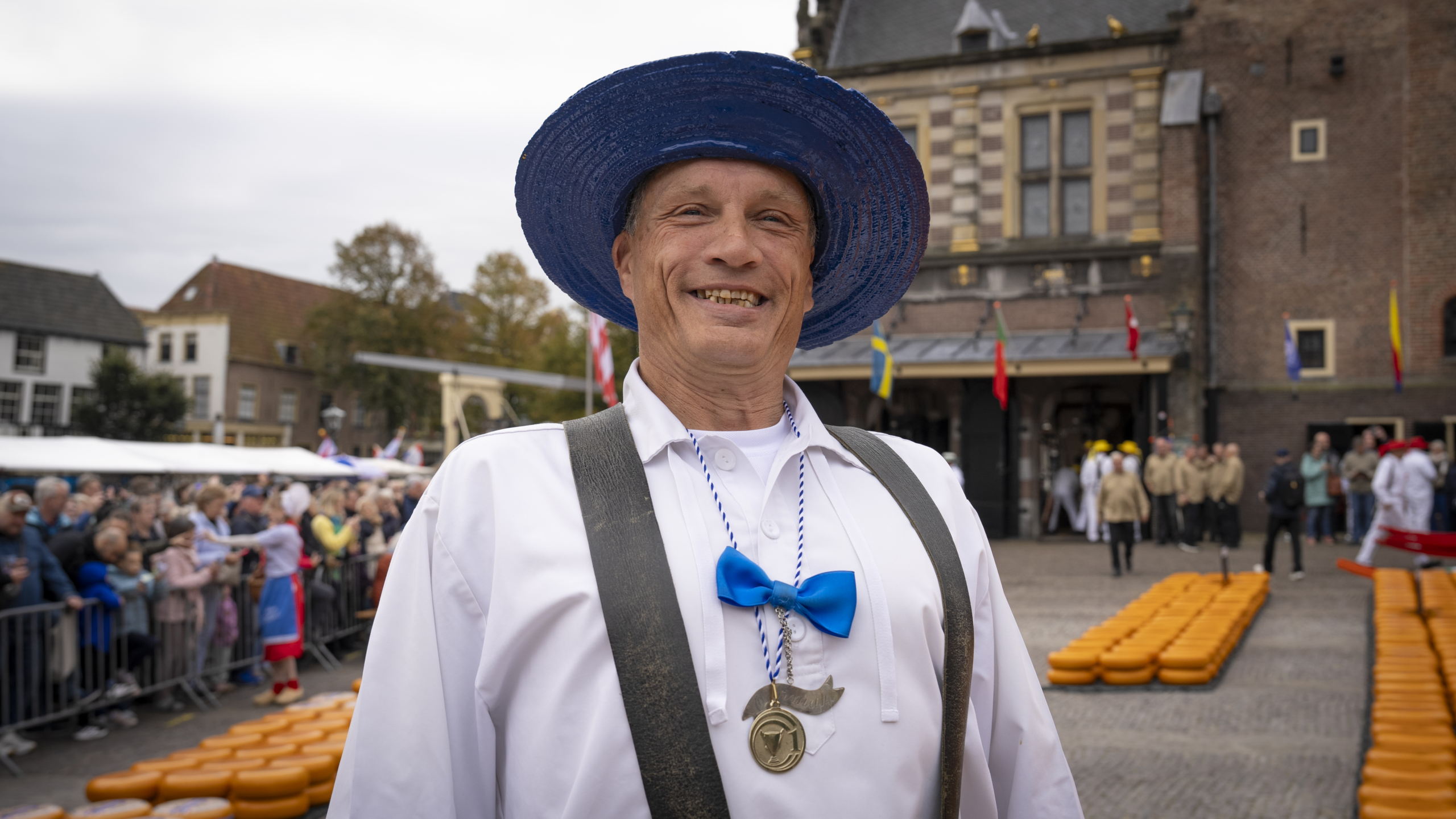 Man in traditional Dutch attire with a blue hat and bow tie, smiling at a cheese market scene, with rows of cheese wheels and a crowd in the background.