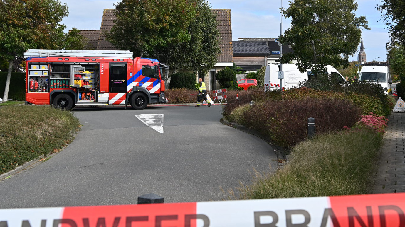 Een brandweerwagen staat op een weg voor een woonhuis, met apparatuur en gereedschap zichtbaar in open zijpanelen. Op de achtergrond zijn enkele bomen, andere voertuigen en een huis zichtbaar, terwijl een veiligheidslint met de tekst 'BRANDWACHT' op de voorgrond hangt.