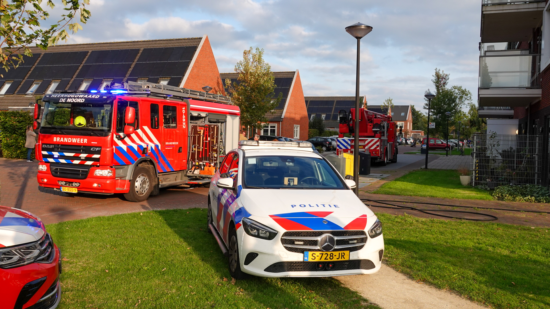 Een brandweerwagen en een politiewagen staan geparkeerd in een woonwijk naast rode huizen met zonnepanelen en groene voortuinen, terwijl mensen in de achtergrond staan te kijken.