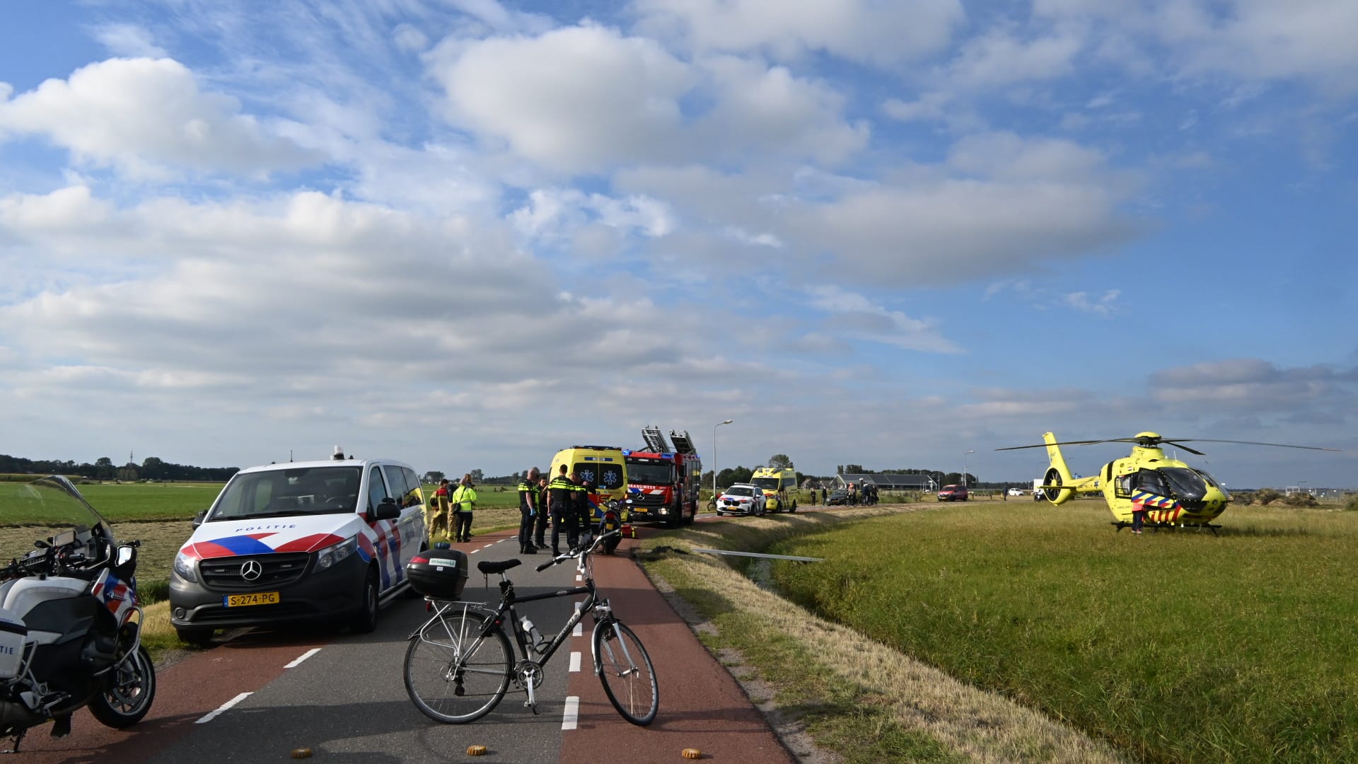 Hulpdiensten op een weg met een politieauto, ambulance, brandweerwagen en helikopter, omringd door open grasland en een heldere hemel.
