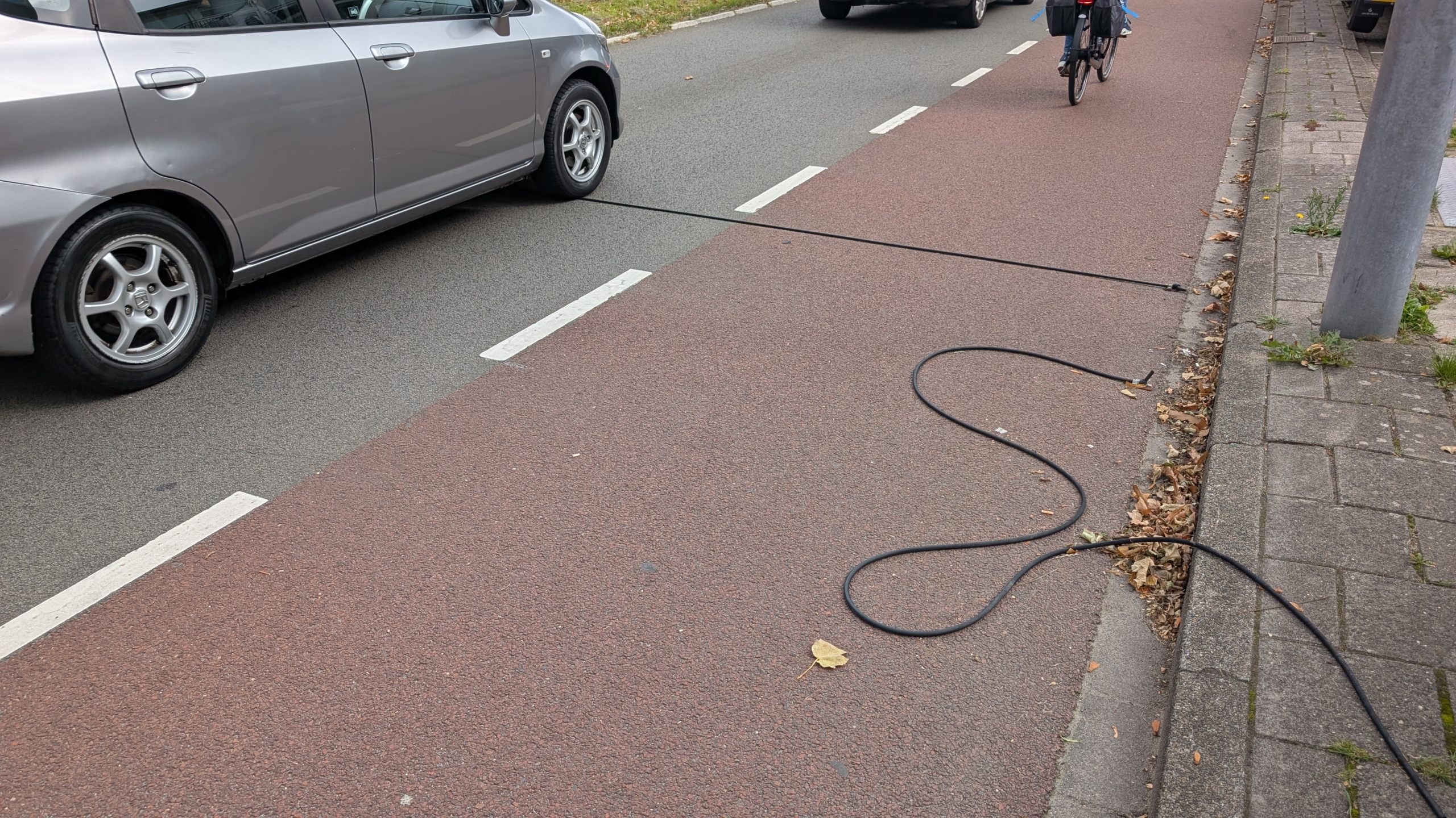 Een auto en een fietser passeren een dunne zwarte kabel die over een weg en fietspad is gespannen om verkeer te tellen, met bladeren langs de stoeprand.
