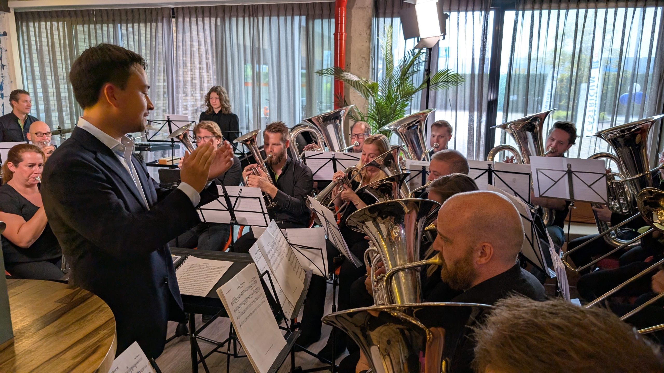 Dirigent leidt een brassband met muzikanten die koperblaasinstrumenten bespelen in een ruimte met gordijnen en kamerplanten.