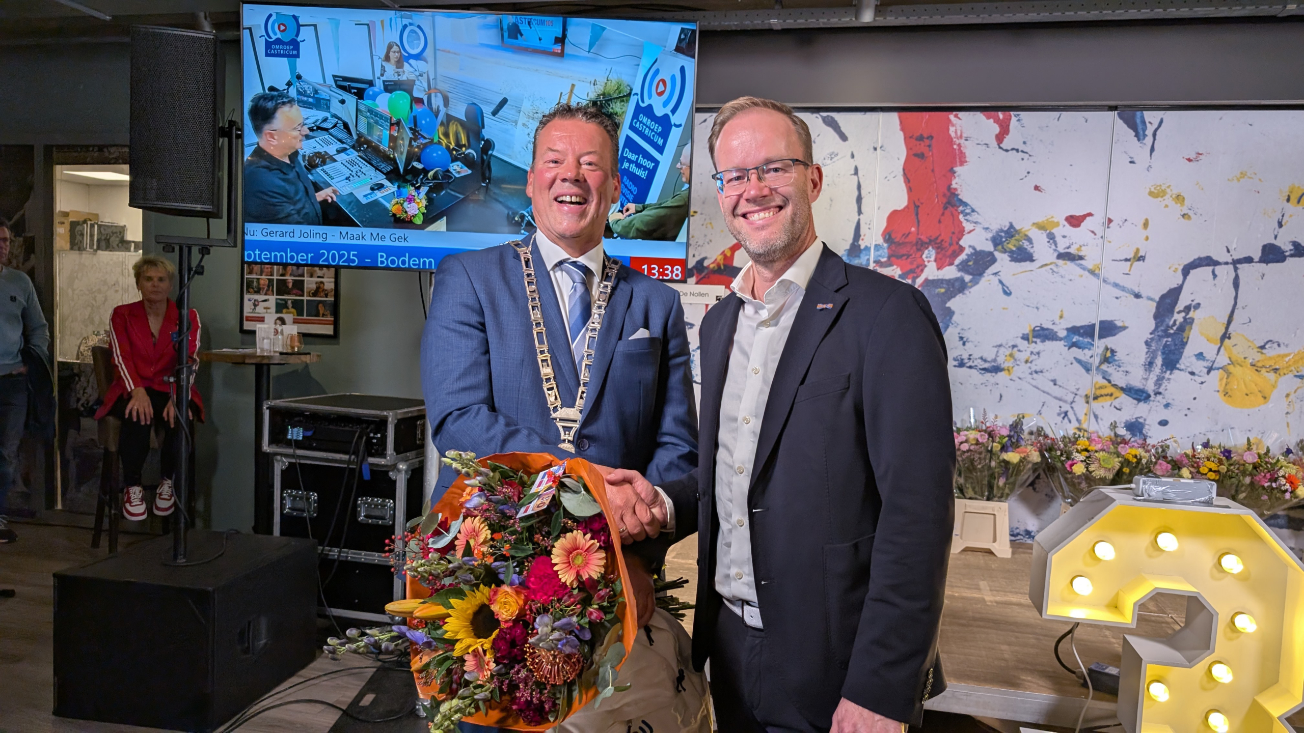 Twee mannen schudden handen terwijl een van hen een bloemenboeket vasthoudt; op de achtergrond is een televisiescherm te zien met een afbeelding van een radiostudeo, en er zijn mensen in de kamer.