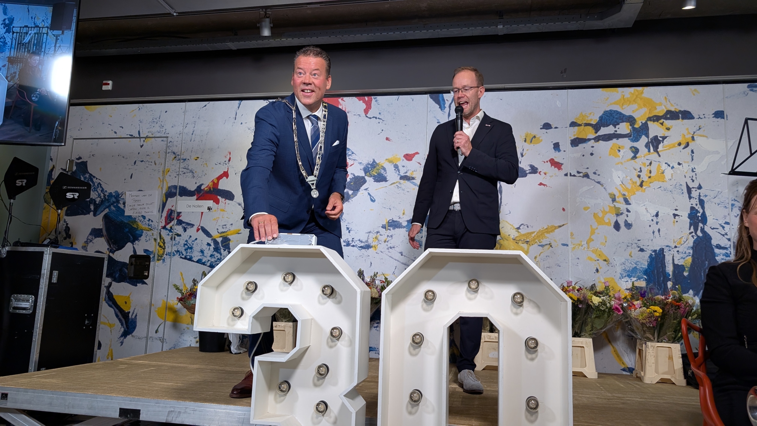 Twee mannen in pak staan op een podium met een feestelijke achtergrond; een van hen houdt een microfoon vast terwijl de ander lacht naast grote verlichte cijfers "20". Op de achtergrond staan bloemen.