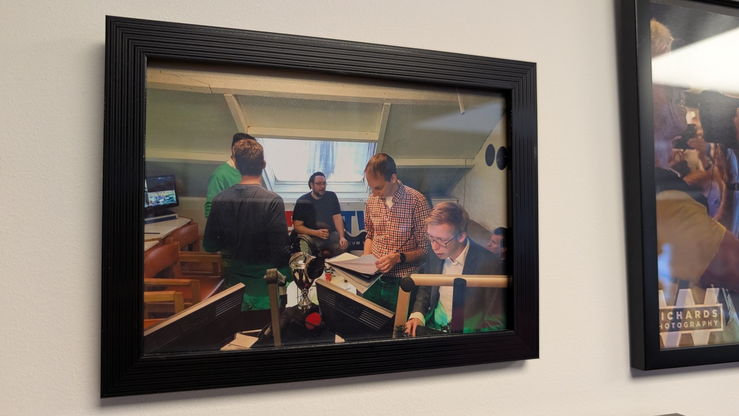 Ingelijste foto van een groep mannen in een kantooromgeving, werkend aan computers met papieren en documenten om hen heen.