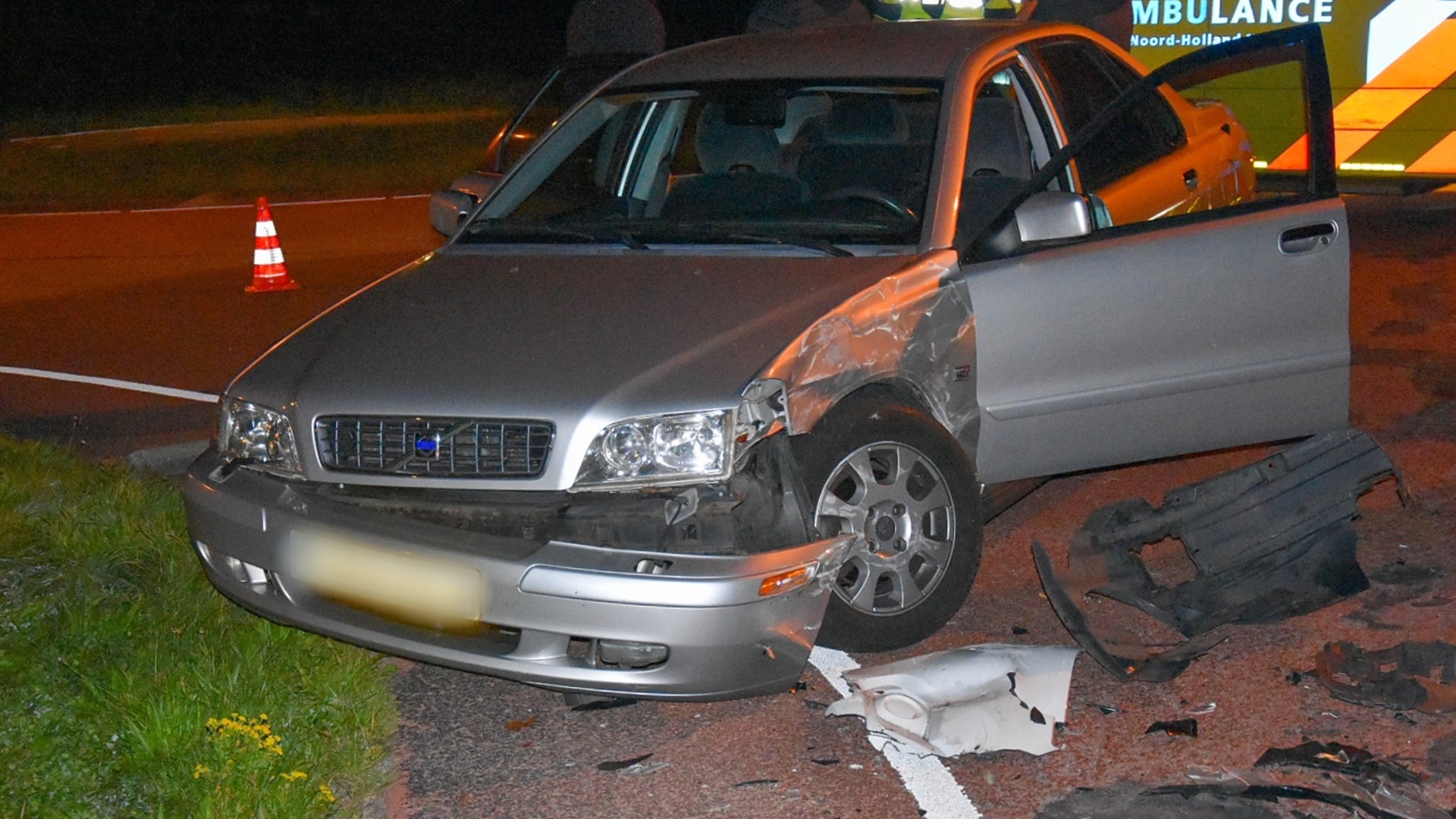 De voorzijde van een lichtgrijze auto met schade aan de bumper en zijkant, geparkeerd langs een verlichte weg met een oranje verkeerskegel. Op de achtergrond staat een ambulance geparkeerd.