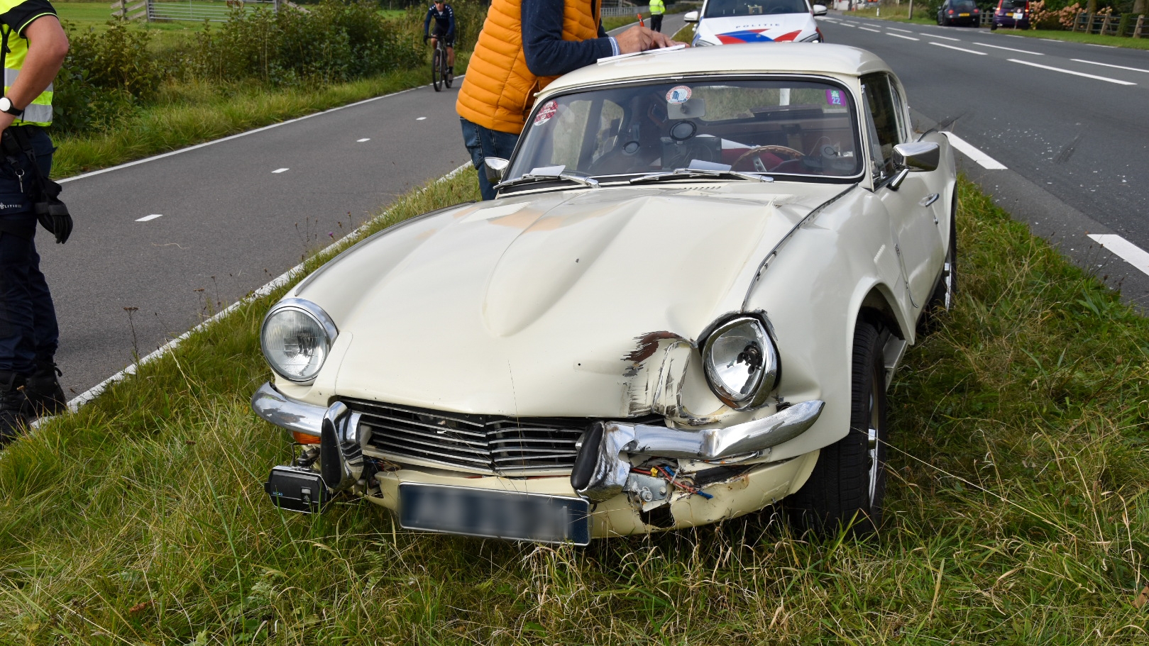 Een witte klassiek ogende auto met schade aan de voorzijde staat langs de kant van een weg, met een persoon in een oranje jas die ernaast staat te schrijven en een politieagent in de buurt.