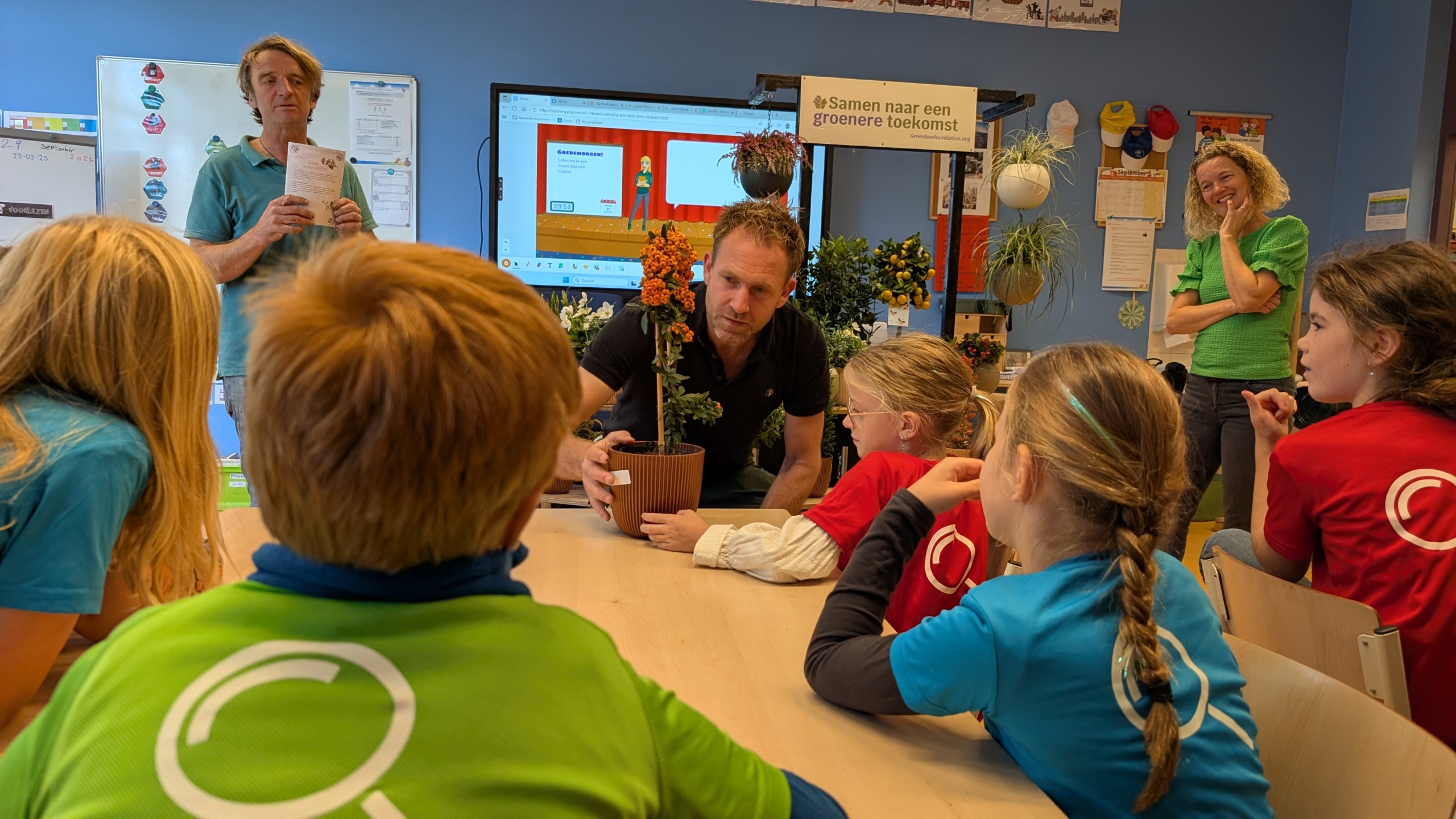 Een man demonstreert een plant in een bloempot aan een groep kinderen in een klaslokaal, met op de achtergrond een scherm en een bord met de tekst 'Samen naar een groenere toekomst'.