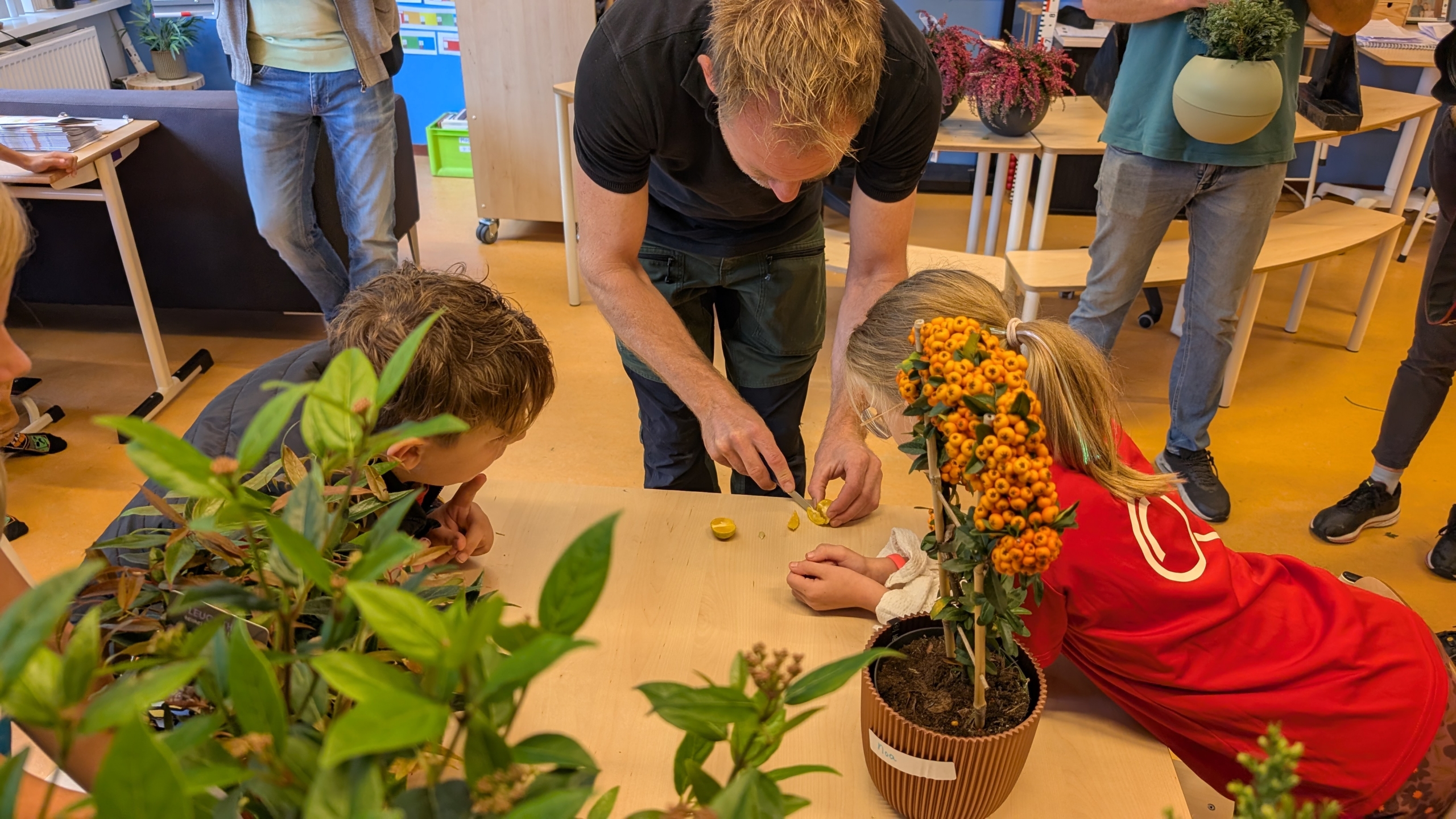 Volwassenen en kinderen verzamelen zich rond een tafel in een klaslokaal, terwijl een man uitleg geeft over iets dat hij snijdt met een mes. Er staan planten en bloemen op de tafel.