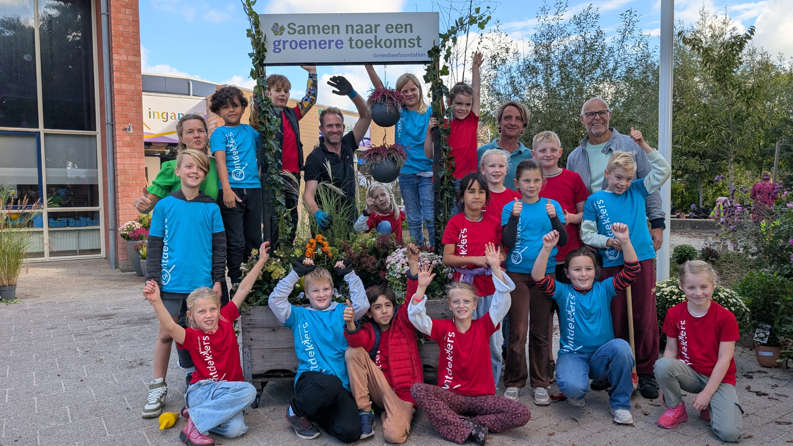Groep kinderen en volwassenen poseren enthousiast voor een plantenbak met kleurrijke bloemen en een bord met de tekst 'Samen naar een groenere toekomst'.