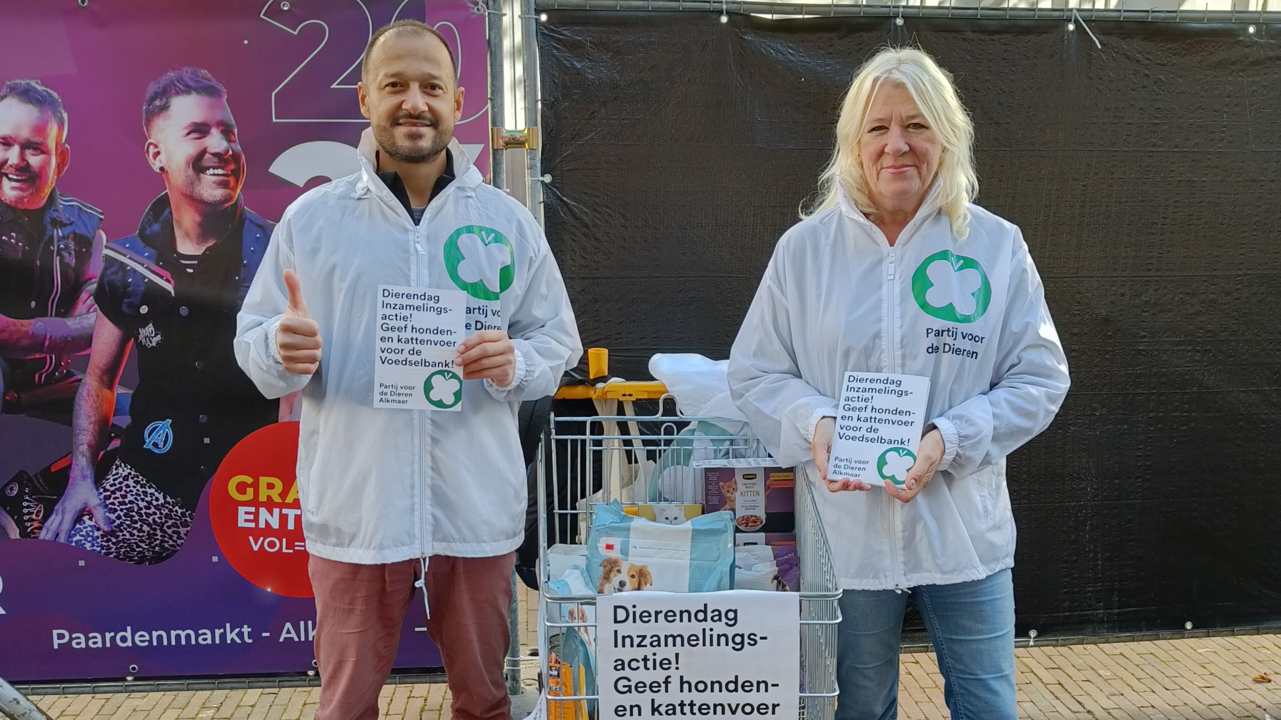 Twee mensen, gekleed in witte jassen met het logo van 'Partij voor de Dieren', staan naast een winkelwagen gevuld met dierenvoedsel. Ze houden borden vast met de tekst: 'Dierendag Inzamelingsactie! Geef honden- en kattenvoer voor de voedselbank!' Op de achtergrond is een kleurrijke poster met afbeeldingen van mensen zichtbaar.