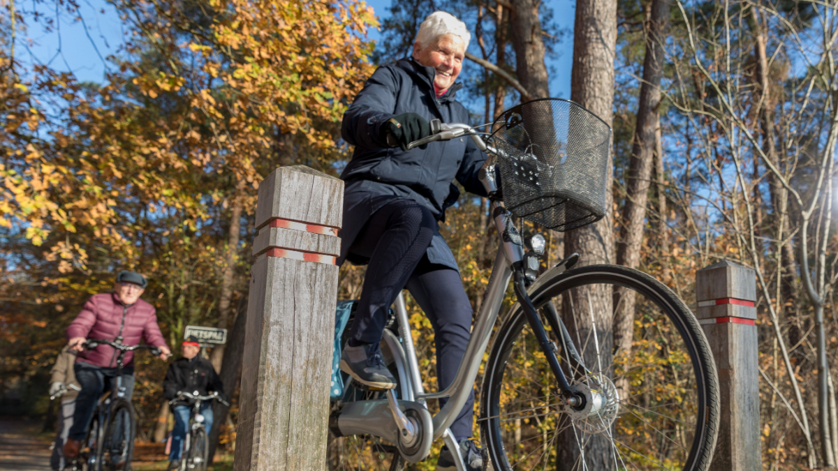 Oudere vrouw fietst met plezier door een bosrijk gebied in de herfst, gevolgd door andere fietsers.