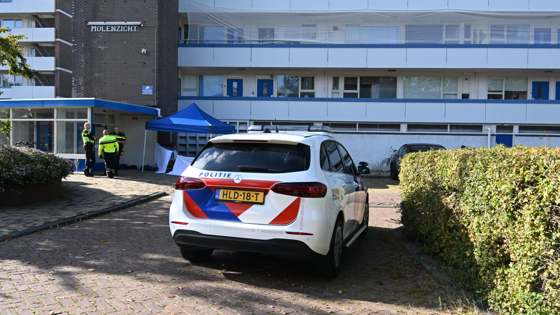 Een politieauto en drie agenten voor een gebouw genaamd 'Molenzicht'. Er is een blauwe tent opgezet bij de ingang van het gebouw.