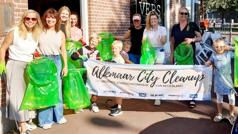Mensen van verschillende leeftijden poseren met groene vuilniszakken en een spandoek met de tekst 'Alkmaar City Cleanup', bij een gebouw met een bakstenen gevel in de zon.