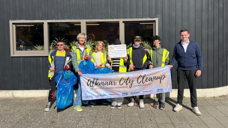Een groep van zes mensen poseert voor een gebouw met een houten gevel, allen gekleed in gele veiligheidsvesten. Ze houden vuilniszakken en afvalgrijpers vast. Voor hen hangt een groot spandoek met de tekst 'Alkmaar City Cleanup'. De lucht is zonnig en blauw.