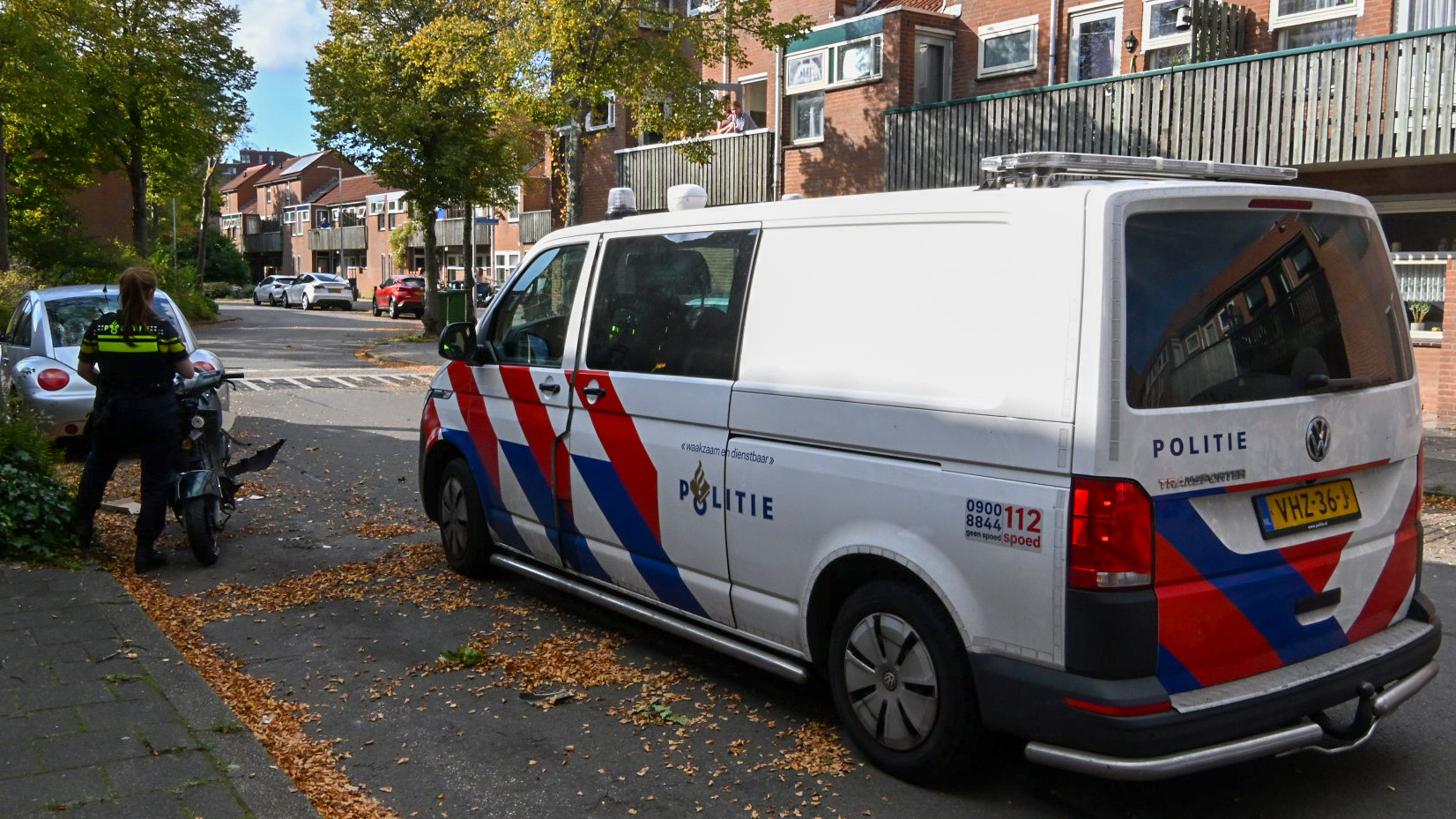 Een Nederlandse politieauto staat geparkeerd naast een agent die bij een scooter staat op een stedelijke straat. Op de achtergrond zijn woonhuizen en bomen zichtbaar.