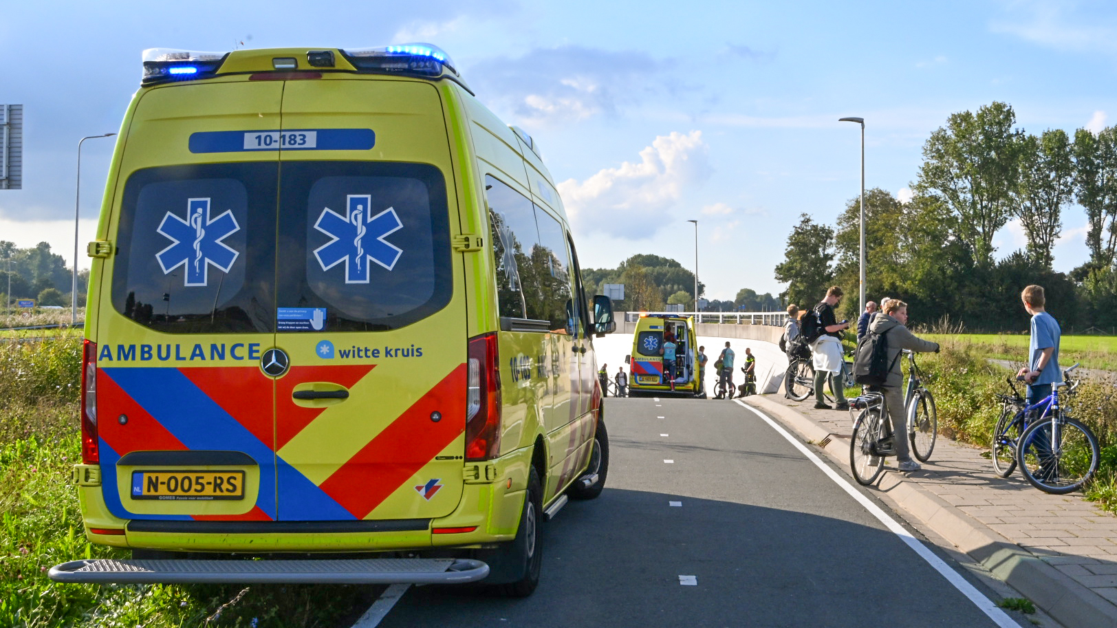 Twee ambulances staan geparkeerd langs de weg met blauwe zwaailichten aan, omringd door mensen met fietsen. De omgeving is groen en bebost.