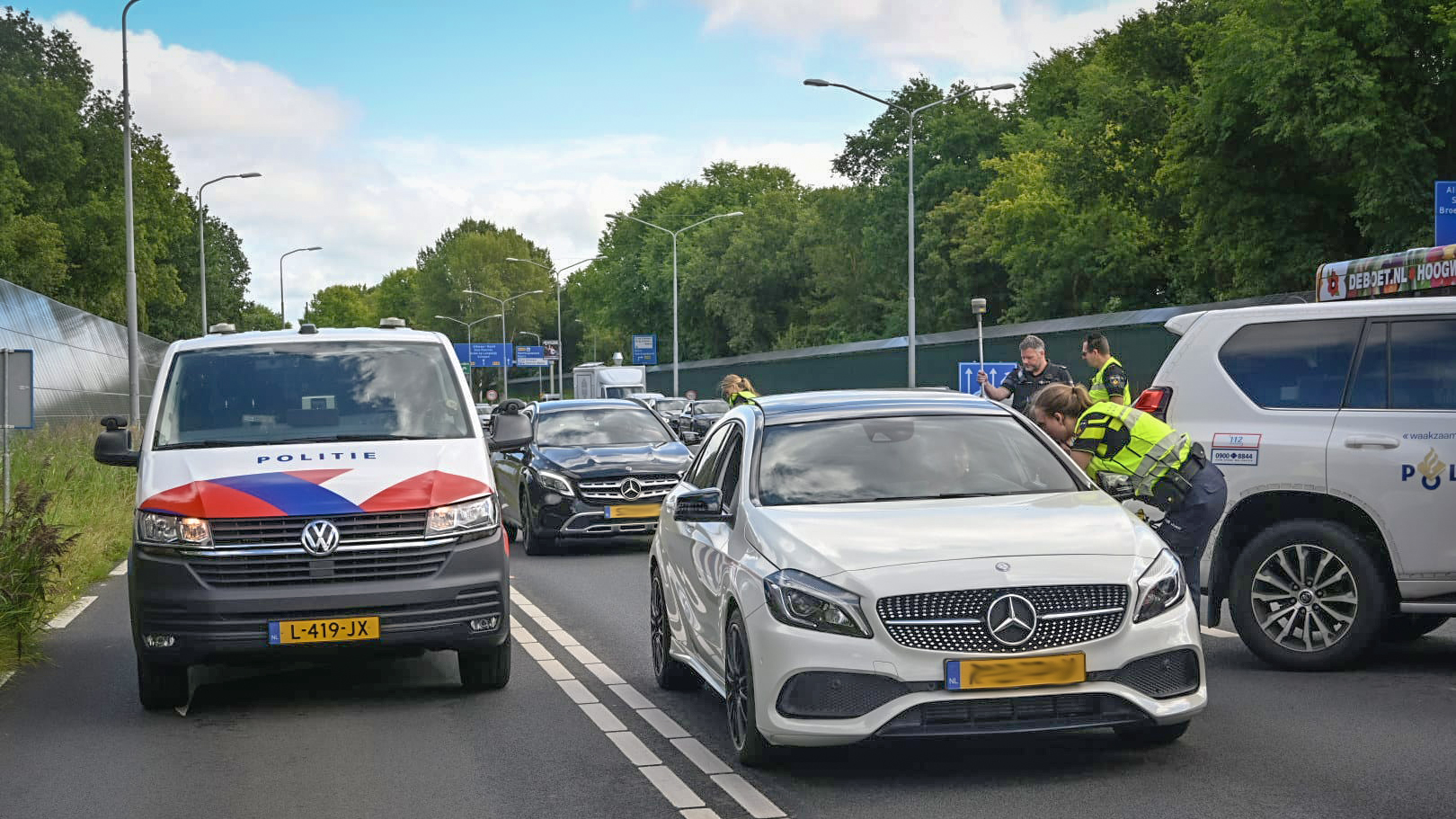 Politieagenten controleren voertuigen op een weg met meerdere rijbanen, waaronder een witte Mercedes en een Volkswagen politiebus in een stedelijke omgeving met veel groen.