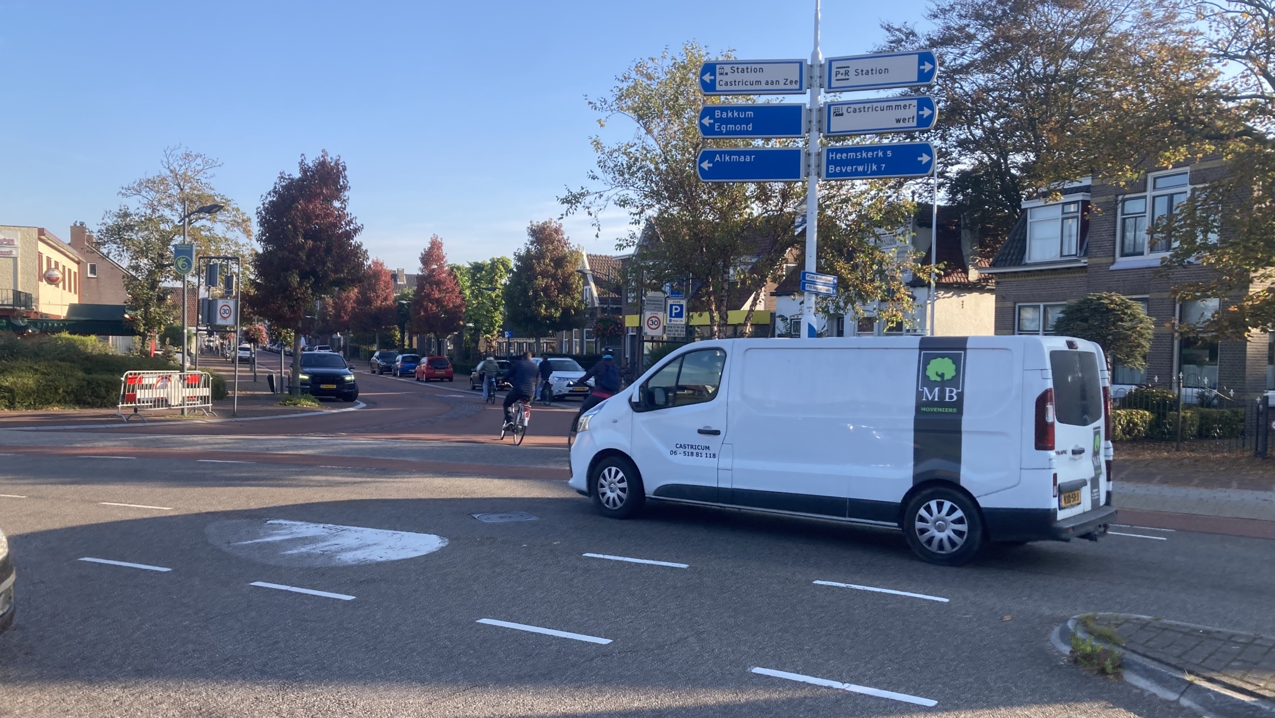 Kruispunt met wegwijzers en verkeer in een Nederlandse woonwijk; een witte bestelbus rijdt op de voorgrond, terwijl mensen op een fiets passeren onder een heldere blauwe hemel.