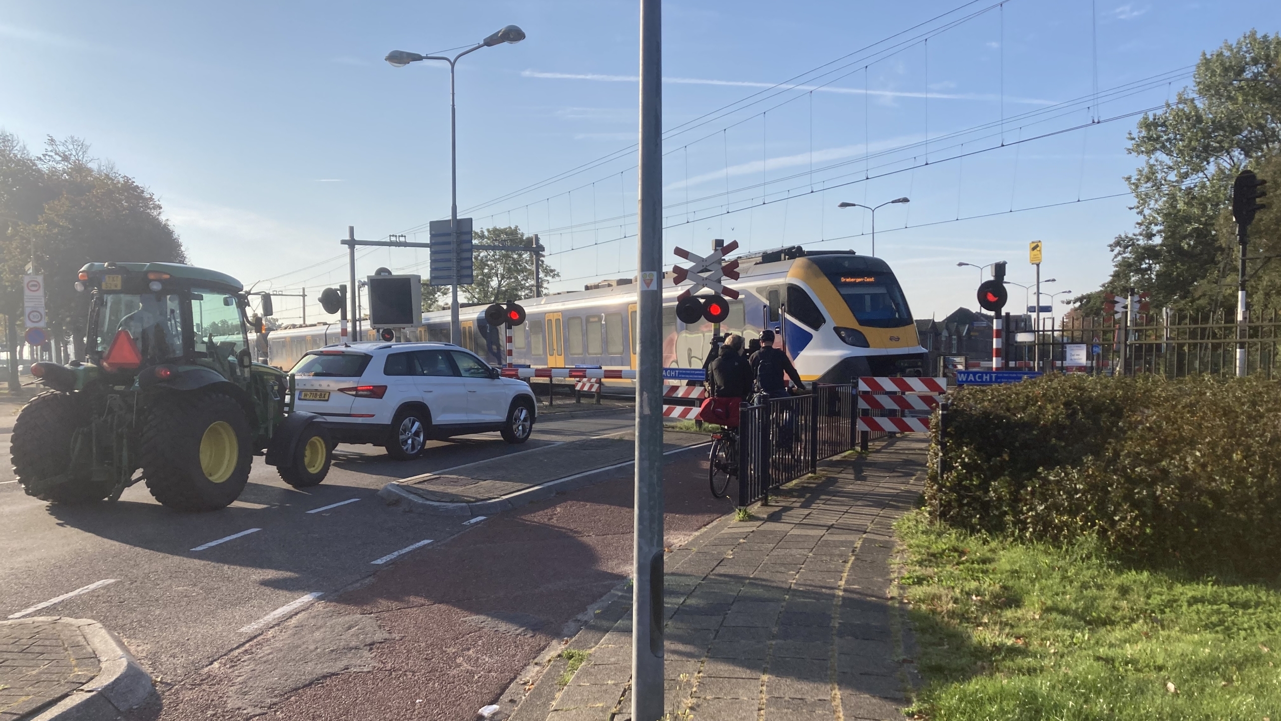 Een witte auto en een tractor wachten bij een gesloten spoorwegovergang terwijl een trein passeert, naast een fietser die ook stilstaat.
