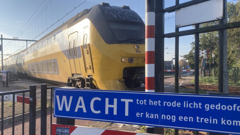 Een geel-grijze trein rijdt langs een spoorwegovergang met een blauw bord waarop 'Wacht tot het rode licht gedoofd is, er kan nog een trein komen' staat geschreven.