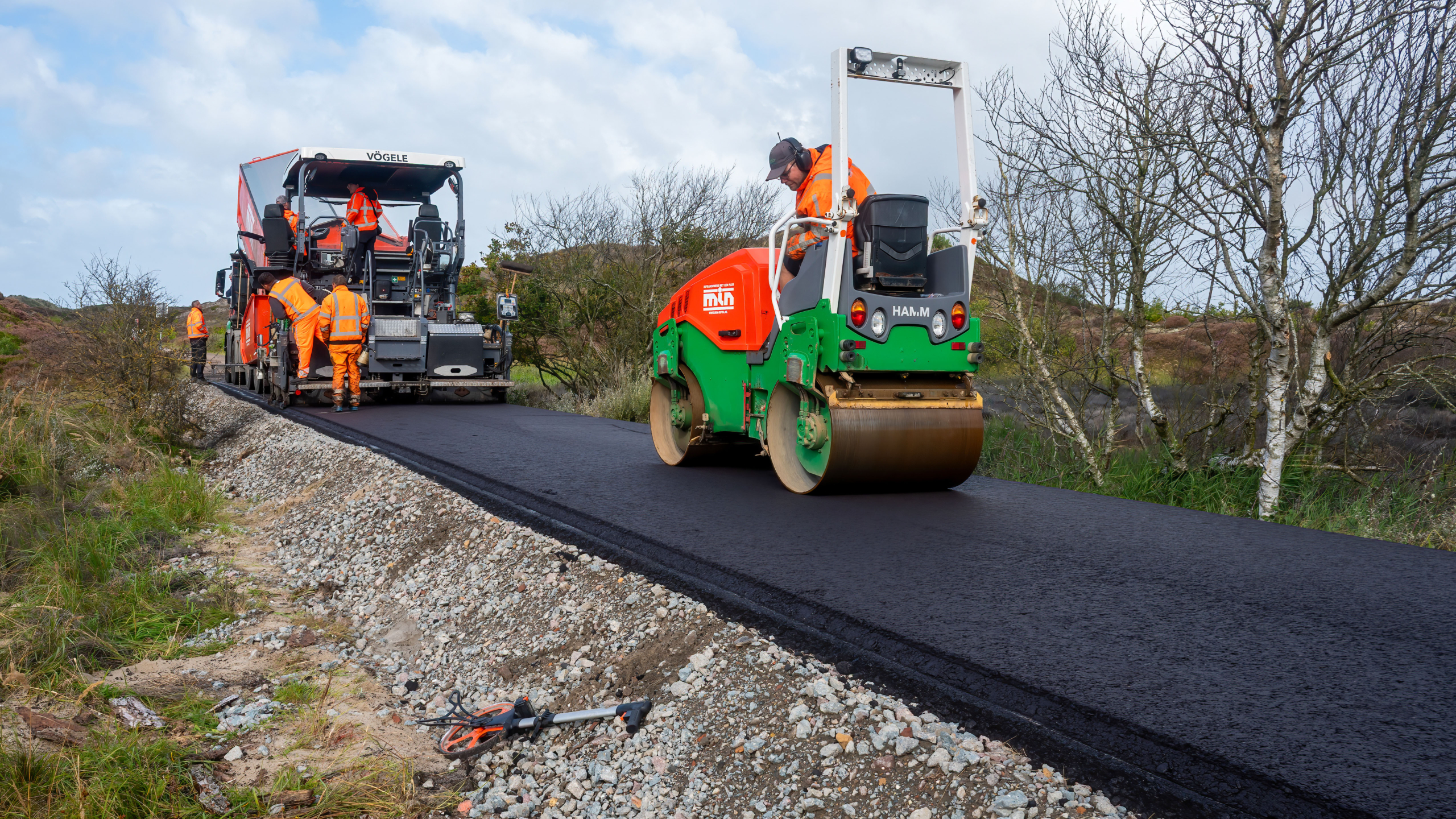 Werkers in oranje kleding leggen een nieuwe asfaltweg aan met een walswagen en een asfalteermachine, omringd door een natuurlijke omgeving met bomen en struiken.