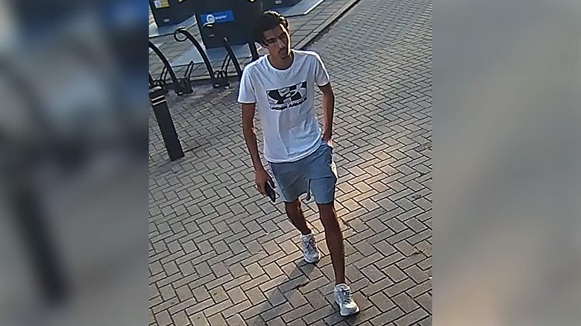 Man met witte T-shirt en korte broek loopt op straat, zijn hand in de zak en telefoon in de andere hand.
