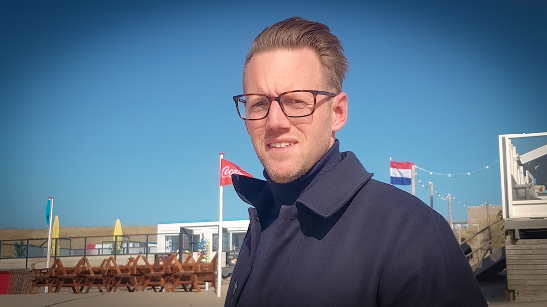 Man met bril en blauwe jas in een buitenomgeving met een Nederlandse vlag en strandtent op de achtergrond.