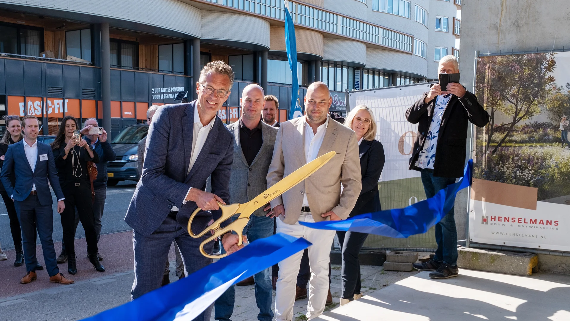 Mensen in nette kleding voeren een lintjesknipceremonie uit met een grote gouden schaar voor een nieuw gebouw, terwijl toeschouwers foto's maken.
