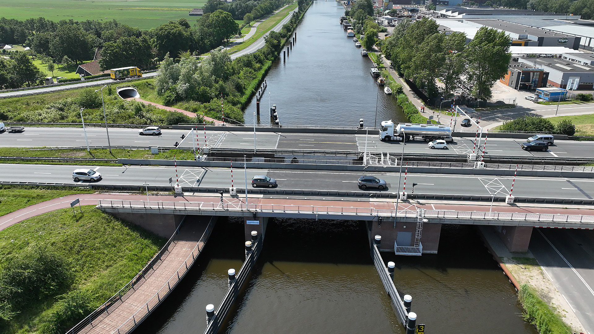Luchtfoto van een brug over een kanaal met verkeer, waaronder auto's en een vrachtwagen. Er zijn wegen en fietspaden naast het water, omgeven door groene velden en gebouwen in de verte.