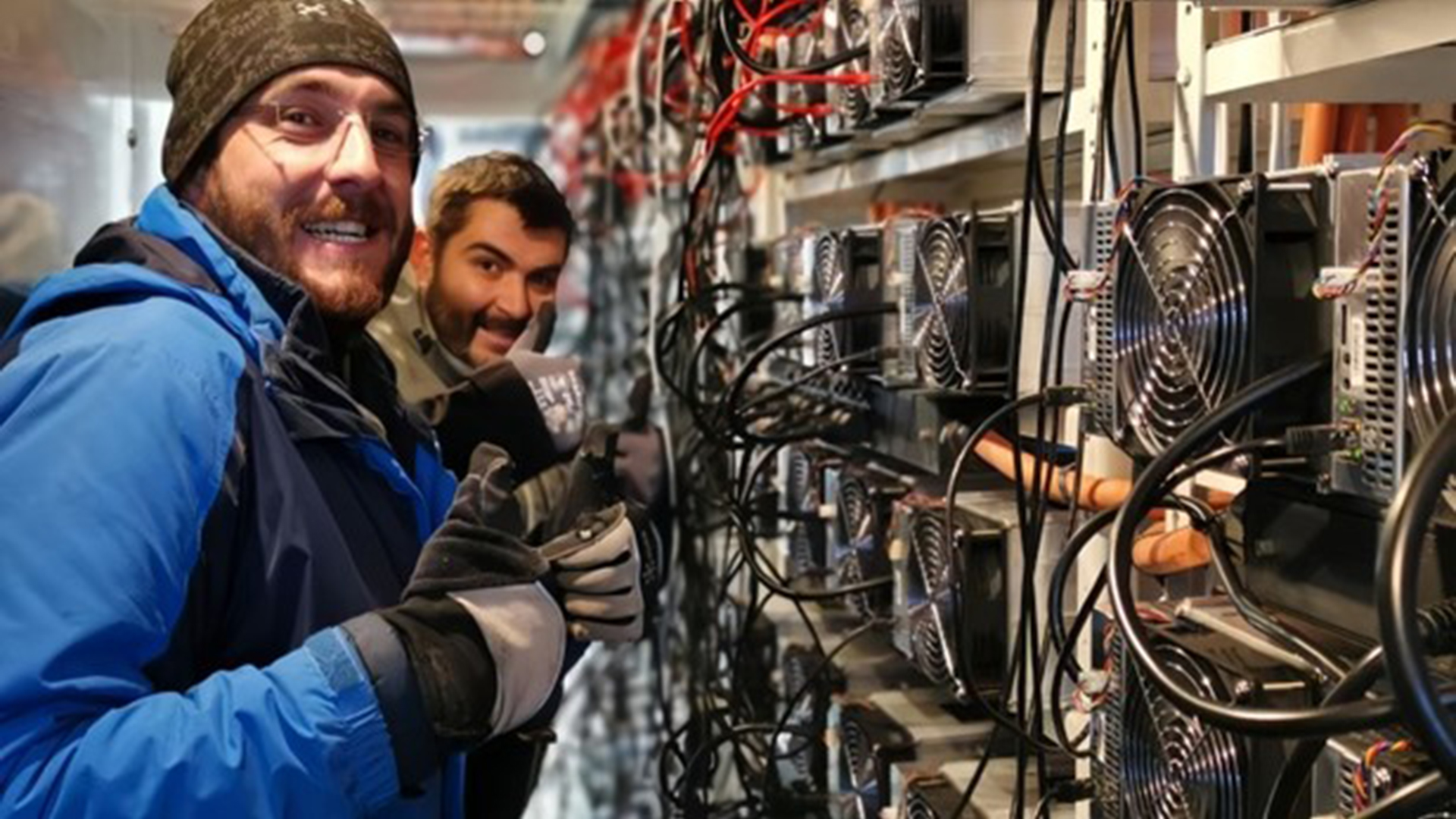 Twee mensen in winterkleding staan lachend voor een rij computerapparatuur met ventilatoren en kabels.