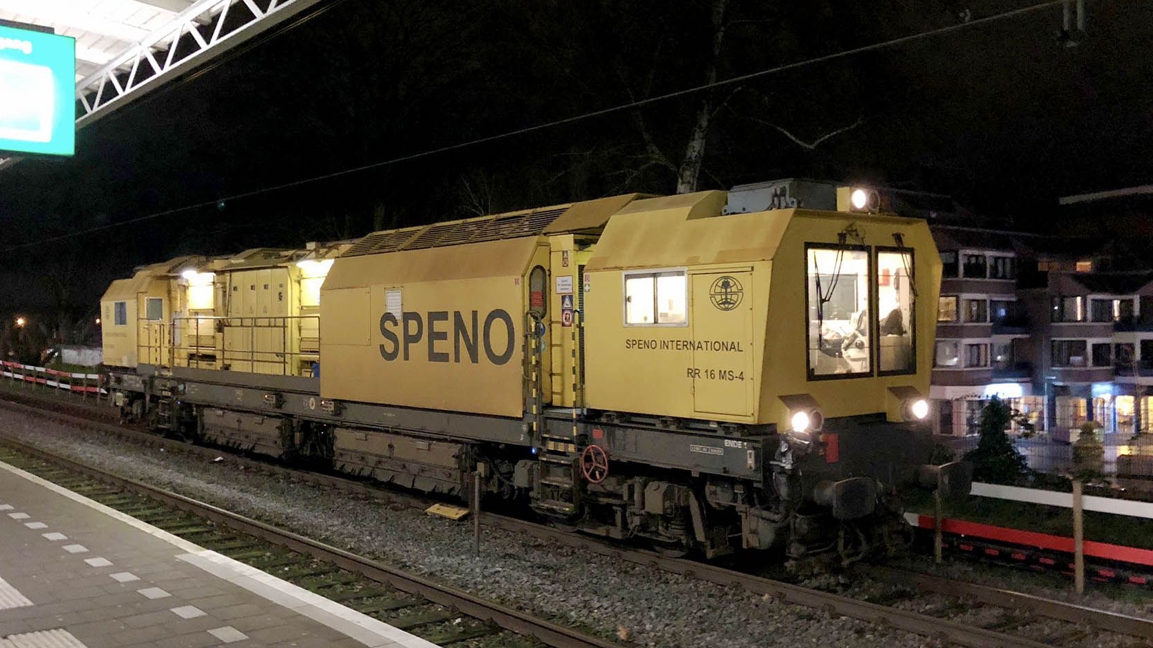 Gele Speno spooronderhoudstrein geparkeerd op een station in de avond.