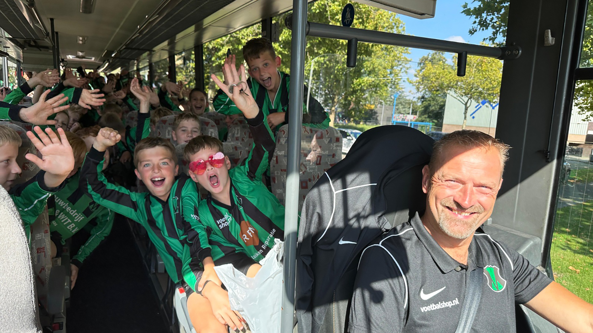 Een groep kinderen in groene en zwarte sportkleding juichen op een bus, met een glimlachende buschauffeur op de voorgrond, terwijl ze naar buiten kijken op een zonnige dag.