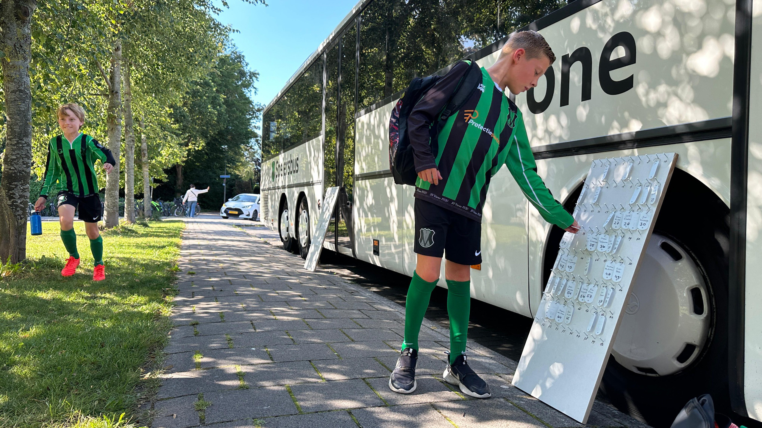 Twee jongens in groene en zwarte voetbaltenues staan bij een bus; de een loopt op het gras terwijl de ander een bord met sleutelhangers beoordeelt.