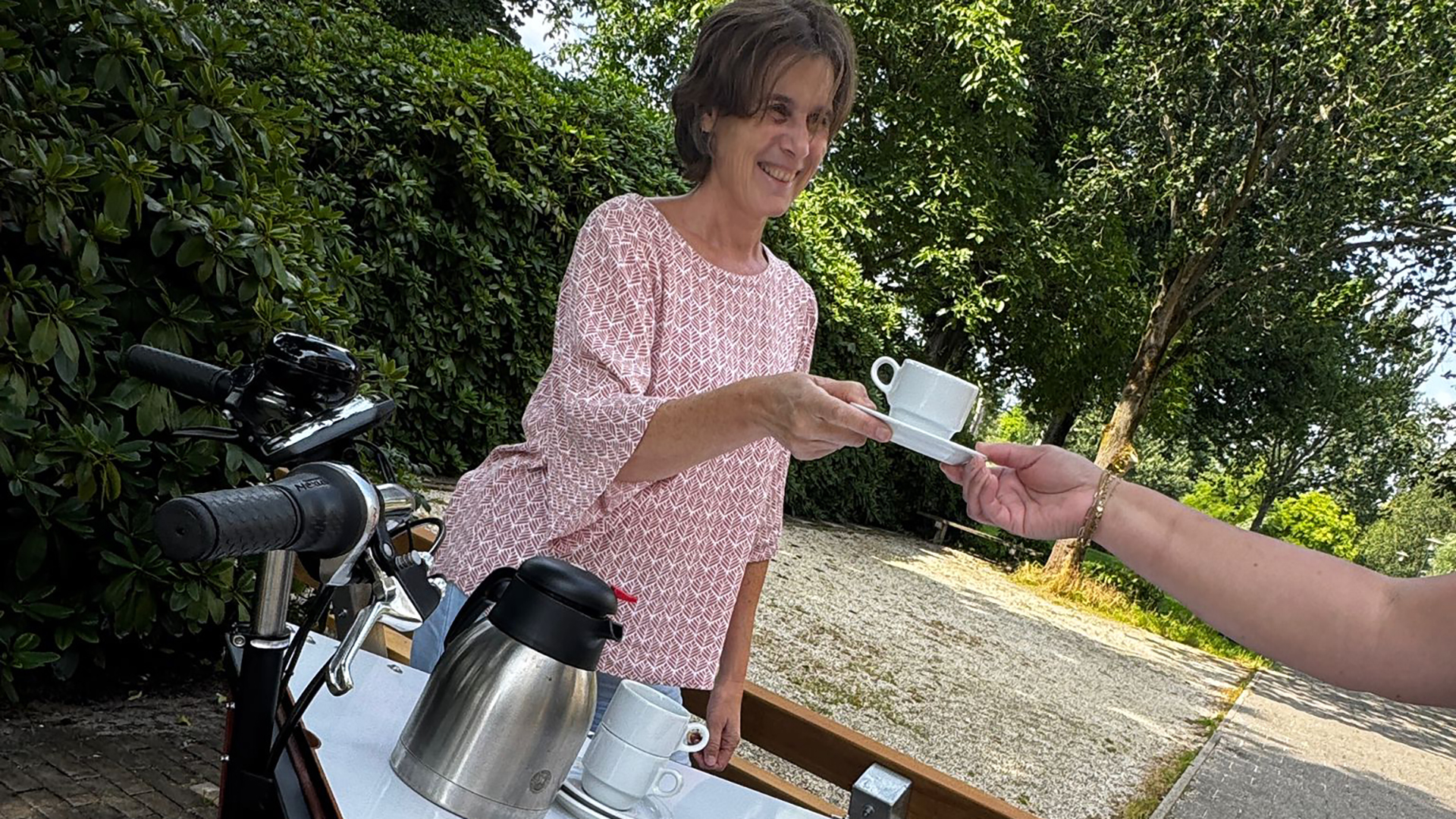 Een vrouw in een roze blouse reikt een kopje koffie aan een persoon op een zonnige dag, met een thermoskan en een fiets op de voorgrond.
