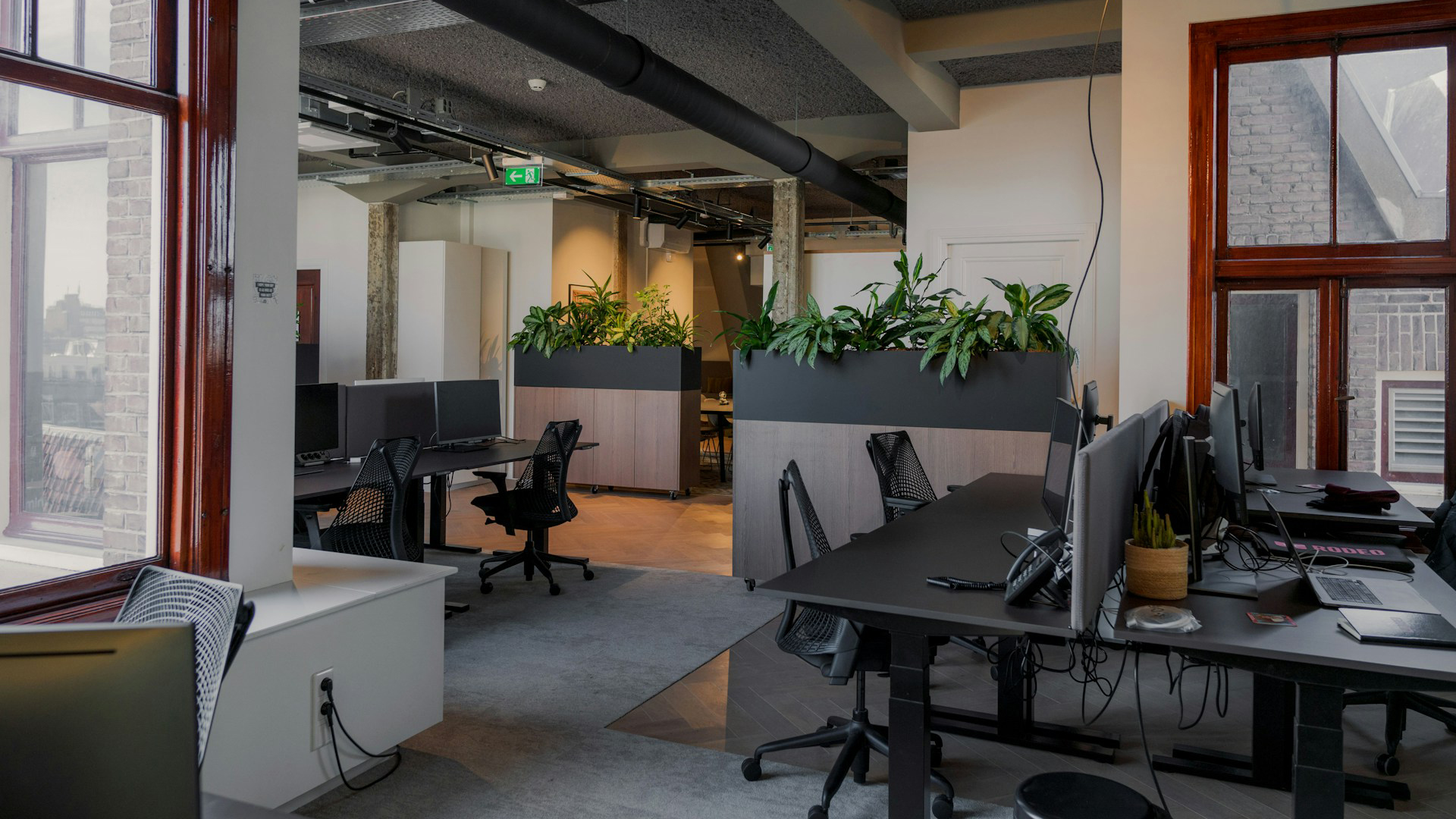 Moderne kantoorruimte met bureaus en stoelen, computermonitoren, en plantenarrangementen als afscherming; grote ramen bieden uitzicht op bakstenen gebouwen buiten.