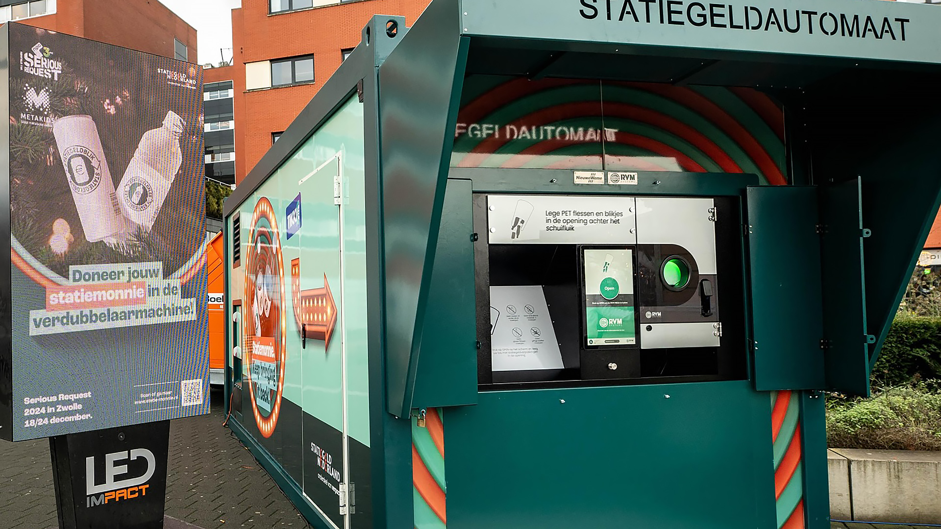 Een statiegeldautomaat naast een LED-scherm met promotie in een stedelijke omgeving. Het scherm toont een advertentie voor het doneren van statiegeld met flessen en blikjes. De automaat heeft instructies voor het inleveren van lege PET-flessen en blikjes.